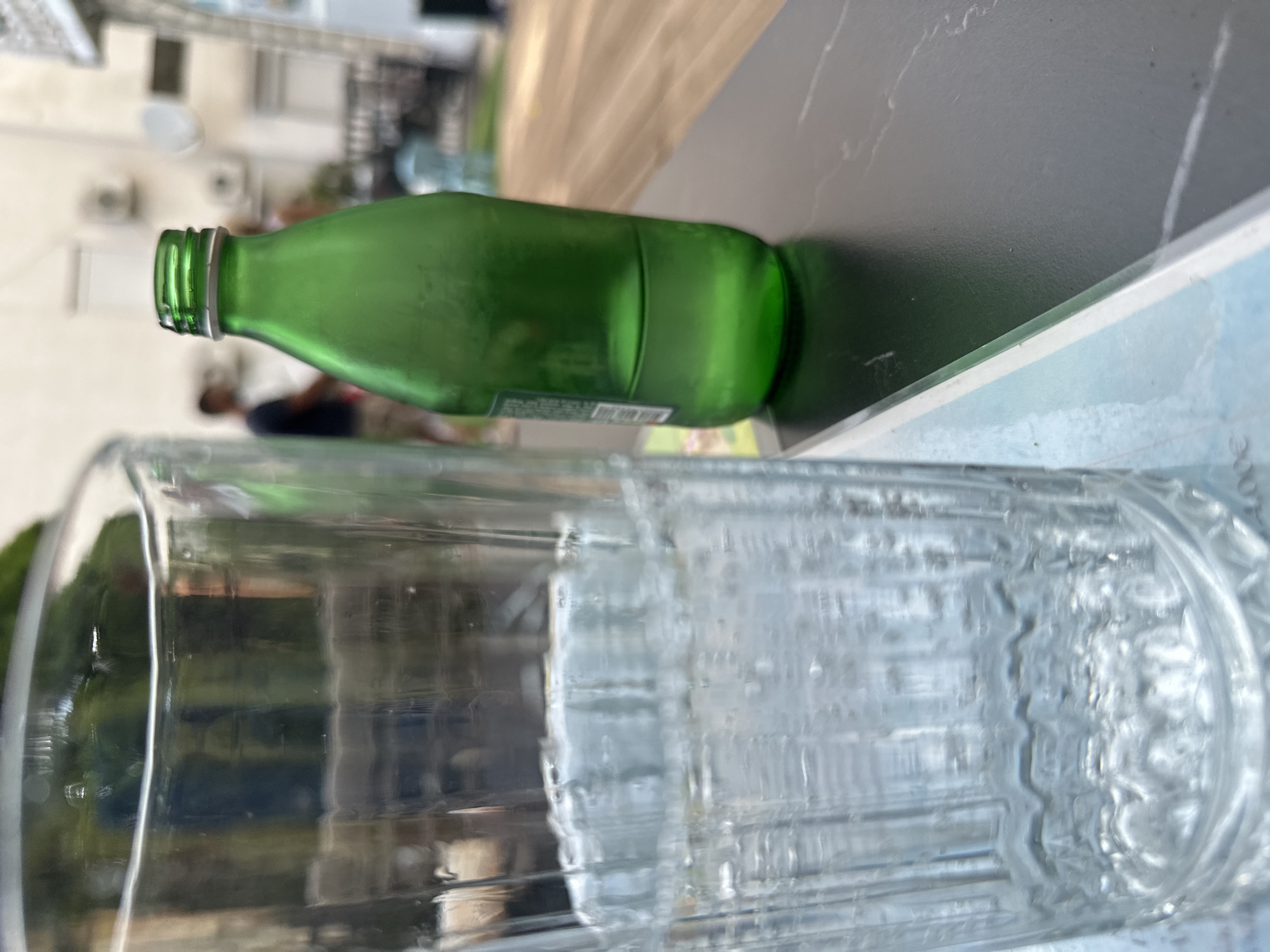 A clear glass filled with water and a green glass bottle containing water are positioned on a dark, marble-patterned table. The glass is on the left and is about half full. The bottle, on the right, is mostly empty, with water filling just the bottom third. In the background, blurred figures of people can be seen, and buildings with air conditioning units are visible. The overall setting appears to be an outdoor cafe or restaurant area, possibly in Budva, Montenegro, given the context provided. The image is taken from a close-up perspective, focusing on the drinks.