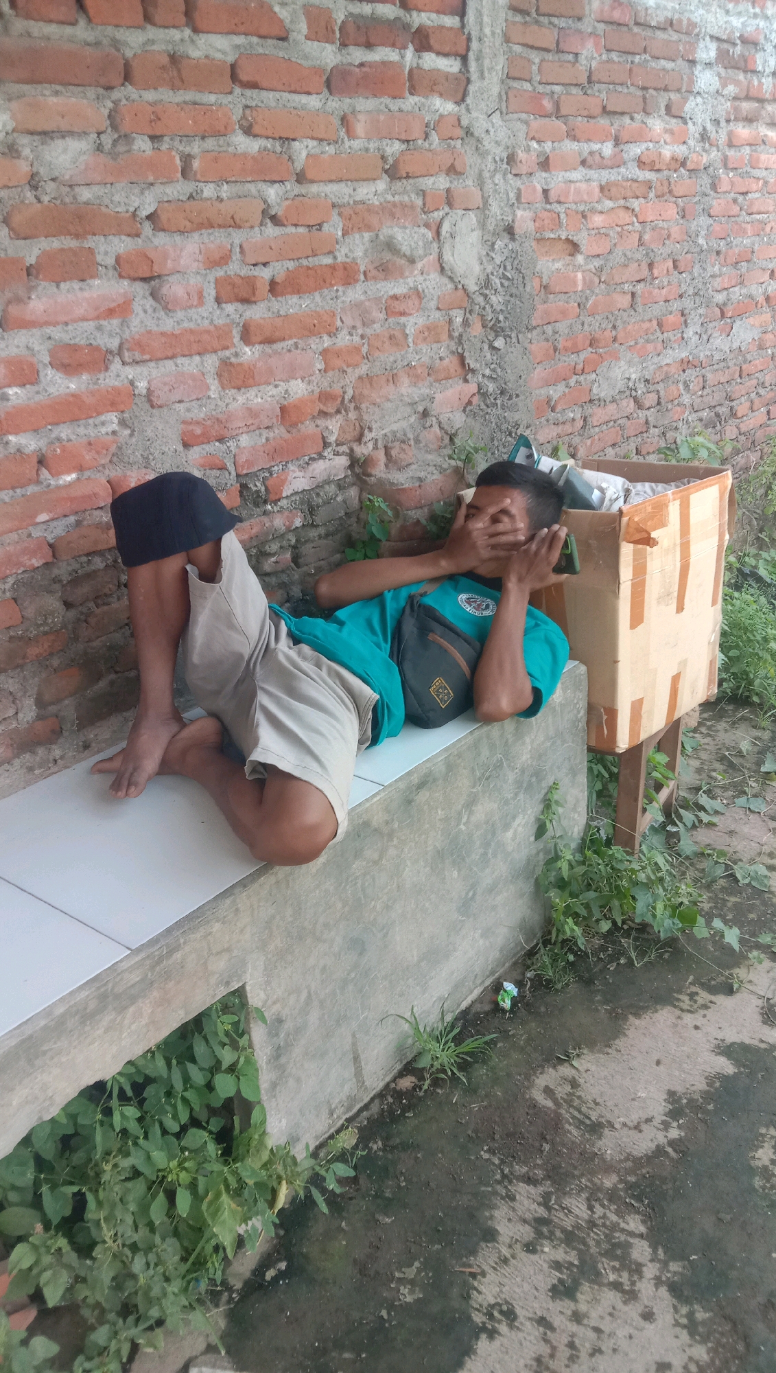 A young male is lying on a concrete ledge in front of a brick wall. He is wearing a teal t-shirt, light brown shorts, and a black cap. He has a black fanny pack slung across his chest. His eyes are covered with his hands. To his right, there is a cardboard box placed on wooden stilts. The ground in front of the ledge is wet and appears to be a mixture of concrete and dirt, with some green vegetation growing around the edges. The scene is likely outdoors, in a part of Karang Anyar, Indonesia.