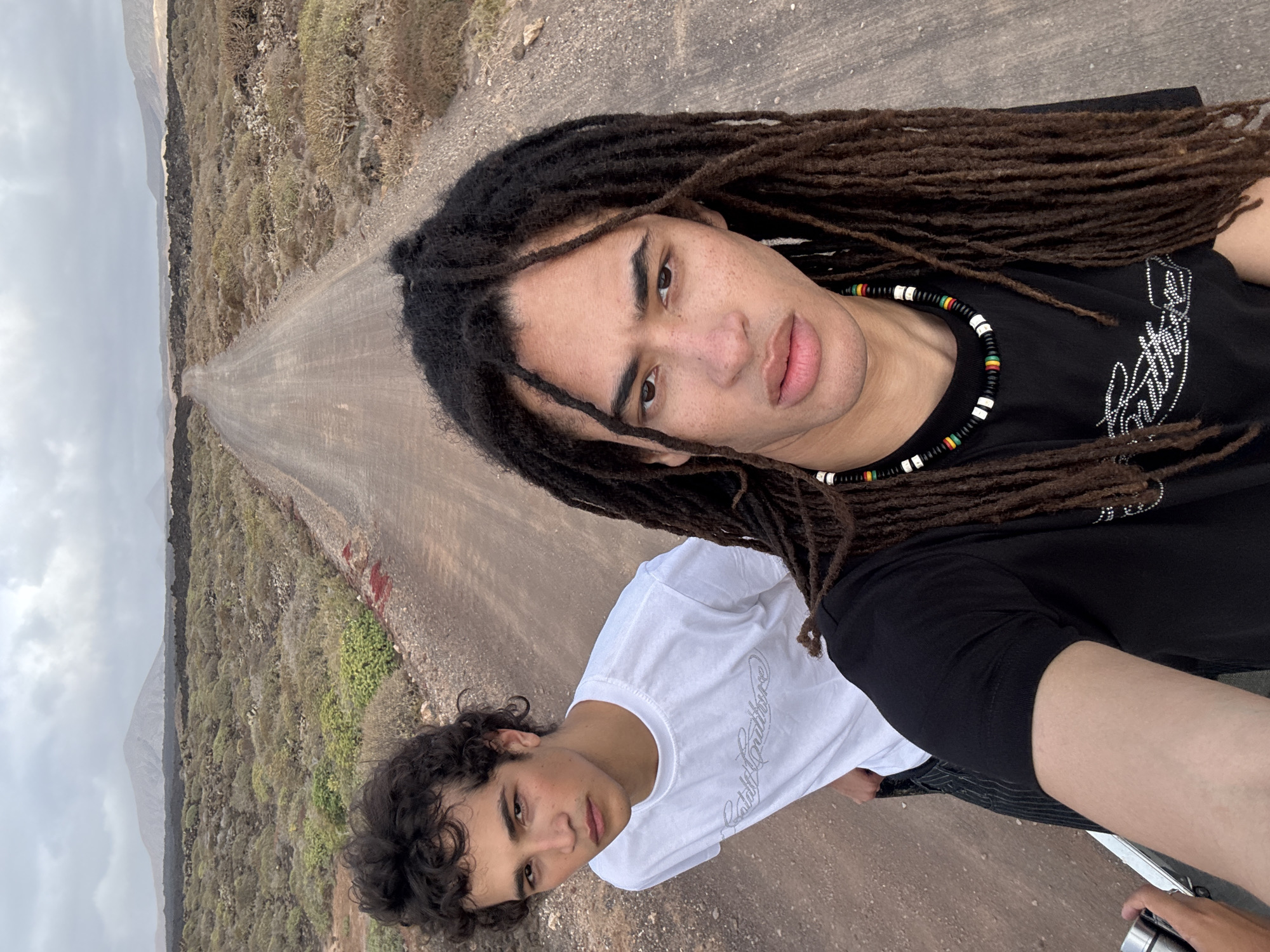 Two young men are pictured in a selfie, against a backdrop of a landscape in Tinajo, Spain. The foreground features the two individuals; the one closer to the camera has dreadlocks, is wearing a black t-shirt and a beaded necklace. The other individual is wearing a white t-shirt. Behind them, there is a long, straight road stretching into the distance, flanked by dry, scrub-covered terrain. The sky is overcast. A hint of distant mountains is visible. The ground surface appears to be arid and rocky.