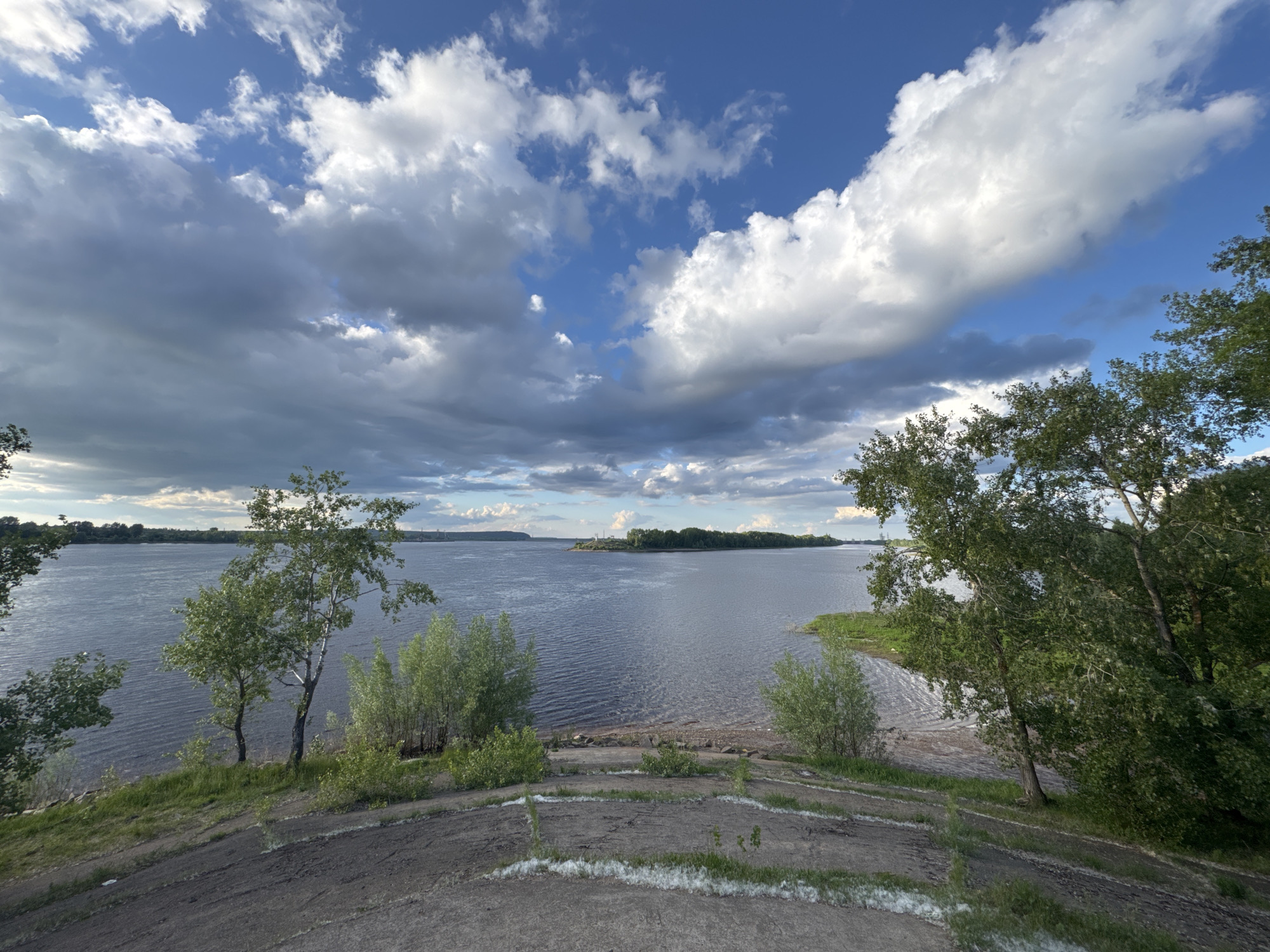 The image depicts a serene landscape scene overlooking a wide river. The foreground is dominated by a sloped, sandy embankment covered with sparse grass and some white fluffy debris, possibly cottonwood fluff. Several young trees and bushes with lush green foliage grow on the embankment, leading down to a pebble-strewn shoreline.
The river itself is calm with gentle ripples, reflecting the dramatic sky. The water appears a deep blue-grey. Across the river, a tree-lined bank forms a distant horizon, broken by a small island with a lighthouse-like structure. The sky is a striking mix of bright blue patches and large, billowing clouds, some dark and heavy with rain, others bright white and illuminated by sunlight.
The viewpoint appears to be from a elevated position on the embankment, looking out over the Kama River in the city of Chaikovsky, Russia. The overall atmosphere is one of natural beauty and tranquility, with the dynamic sky adding a sense of drama to the peaceful river scene. No people are visible in the image.