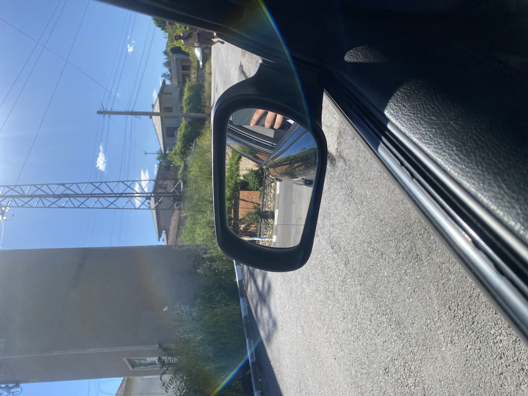 The image captures a scene from inside a vehicle, looking out a side window and into the side-view mirror. The car is on an asphalt road, which appears to be in the city of Sadovets, Bulgaria, evidenced by the distinctive electrical infrastructure and architectural style of the buildings.
In the foreground, the interior of the car is visible, with a textured dark surface on the door. The side-view mirror reflects a portion of the road and the environment behind the car. A hand, presumably belonging to the driver or a passenger, is visible inside the car, holding a smartphone.
Outside the car, the road curves slightly. To the left, a tall, plain concrete structure stands near the roadside, with some vegetation growing around its base. A rainbow-like lens flare arcs across the scene, originating from the left side. Beyond the road, several houses with tiled roofs are visible, nestled amongst green foliage. An electrical pylon with multiple wires stretches across the bright blue sky, a common sight in rural Bulgarian landscapes. In the distance, a shirtless man is walking away from the camera, wearing dark shorts and a band around his forehead or neck. The overall impression is that of a bright, sunny day in a semi-rural Bulgarian setting.