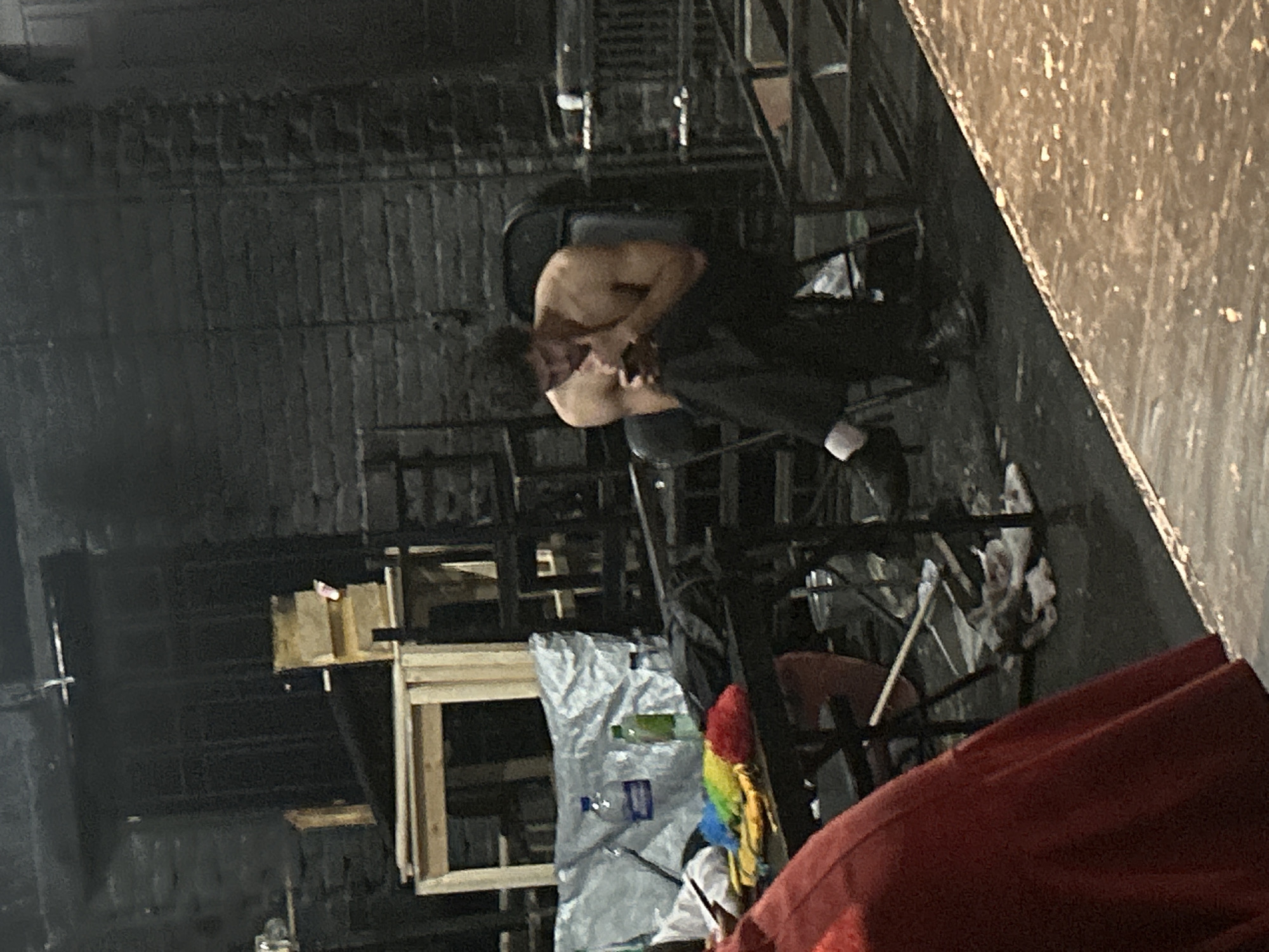 A shirtless man with dark, curly hair sits on a folding chair in a dimly lit, industrial-looking space, likely backstage. He is wearing dark pants and white socks, and his feet are clad in dark shoes. The man is looking down at a smartphone in his hands, his fingers touching the screen. The background is a rough, dark brick wall. In front of him, a table is draped with a large, reflective silver tarp, behind which are two plastic bottles, one clear with a blue label and the other green. Several cleaning supplies, including colorful sponges or brushes, are on the table near the bottles. To the left, a portion of a large red garment is visible, obscuring part of the scene. The floor appears to be concrete or a similar dark, industrial material, with some scattered debris and a visible water stain. The overall impression is of a utilitarian backstage area, possibly a theater or studio.