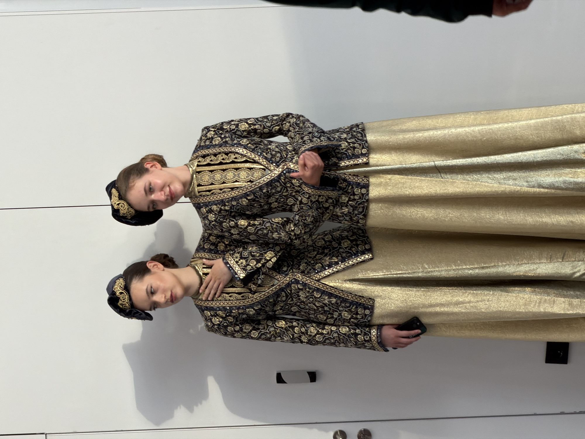 Two young women, appearing to be models, stand in front of a white wall with subtle vertical seams. They are dressed in matching elaborate, traditional outfits. Each outfit consists of a dark blue, fitted jacket with intricate gold embroidery in floral and geometric patterns, a high-necked gold lace overlay, and a floor-length shimmering gold skirt. Both women wear matching dark hats with gold embellishments. The woman on the left has her right hand on her chest, and the woman on the right has her hands clasped in front of her. The woman on the left is holding a black mobile phone in her left hand. A shadow of a profile is cast on the wall behind the woman on the left, appearing as if a face is looking to the right. The overall environment suggests a backstage area or a studio setting.