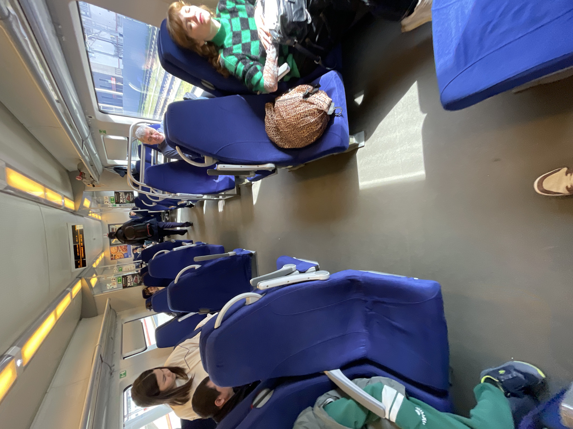The image captures the interior of a train car, likely in Sochi, Russia, based on the Russian text posters visible along the wall. The seats are arranged facing each other in pairs and are upholstered in dark blue fabric. The train is occupied by multiple passengers: two passengers are lying down with their eyes closed and bags on their seats, one wearing a green sweater and the other a leopard print bag. A senior male passenger is looking directly at the camera, other passengers are seen sitting or lying. The lighting in the car is provided by overhead fluorescent lights, some casting a yellow glow, and sunlight filtering in through the windows on the left side of the frame, showing scenery passing by outside. The floor of the train car is dark grey.
