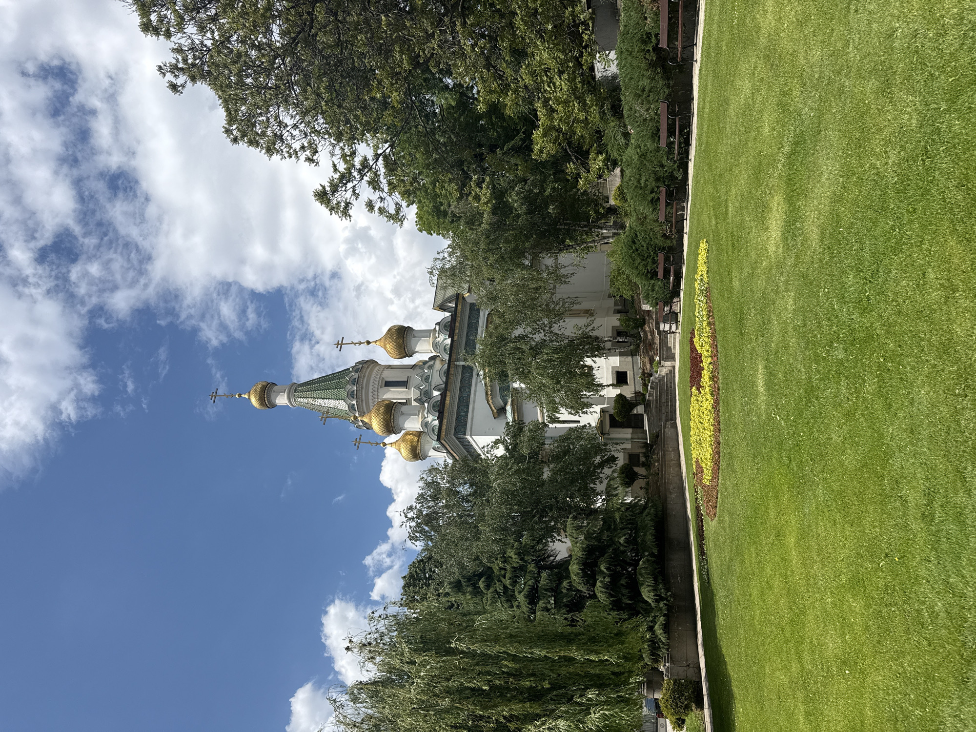 The image shows a partial view of the Russian Church St. Nicholas the Wonderworker in Sofia, Bulgaria. The church features golden domes and a green-tiled roof. The architectural style is distinctive, suggesting Russian Orthodox influence.
The church is partially obscured by trees, and a lawn and flowerbed are visible in the foreground. The sky is blue with scattered clouds.