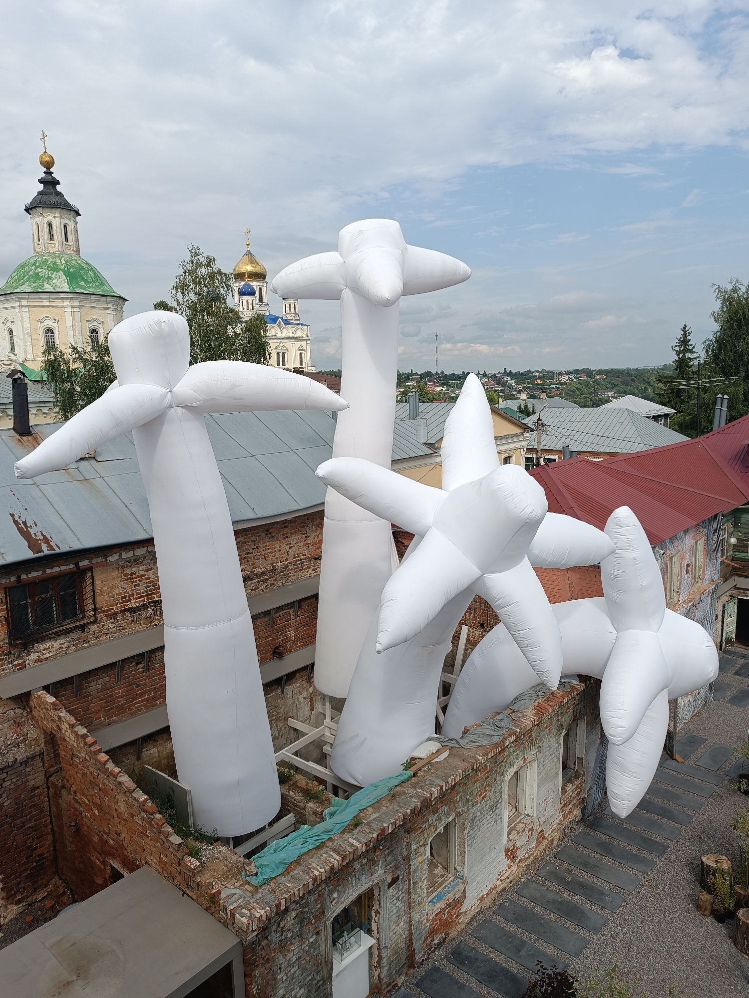The image depicts an outdoor art installation in the city of Yelets, Russia, featuring several large, white, inflatable sculptures resembling abstract figures with outstretched arms or petals. These sculptures are positioned within a courtyard area surrounded by aged brick buildings. Some of the buildings appear to be under renovation or have a distressed aesthetic, with exposed brickwork and empty window frames. In the background, traditional Russian Orthodox churches with distinctive onion domes and crosses are visible, along with a view of the city's rooftops and greenery. The sky is cloudy, suggesting overcast weather. The installation is set against a backdrop that blends contemporary art with the historical architecture of Yelets.