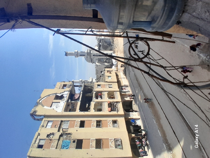 An elevated view captures a street scene in Khan Yunis, Palestinian Territory, marked by significant structural damage.
In the midground, a multi-story, light-colored residential building on the left exhibits severe damage, with large sections of its facade missing, exposing interiors, and visible holes consistent with impact. A blue tarp hangs from a damaged section on an upper floor. At street level in front of this building, clothes are displayed on a rack, suggesting a makeshift stall, with individuals observing or passing by.
Further in the background, a heavily damaged mosque-like structure is visible, with its minaret still standing but visibly scarred, and its main building partially collapsed into a large pile of rubble.
The street, appearing as asphalt, runs through the center, showing some dirt and moisture. Numerous individuals, including children, are walking on the street and sidewalks. On the far right, a partial view of a grey-blue plastic water dispenser bottle and a tangled network of rusty metal rods and wires are prominent in the foreground, suggesting the vantage point is from a damaged building. The sky above is clear and blue.