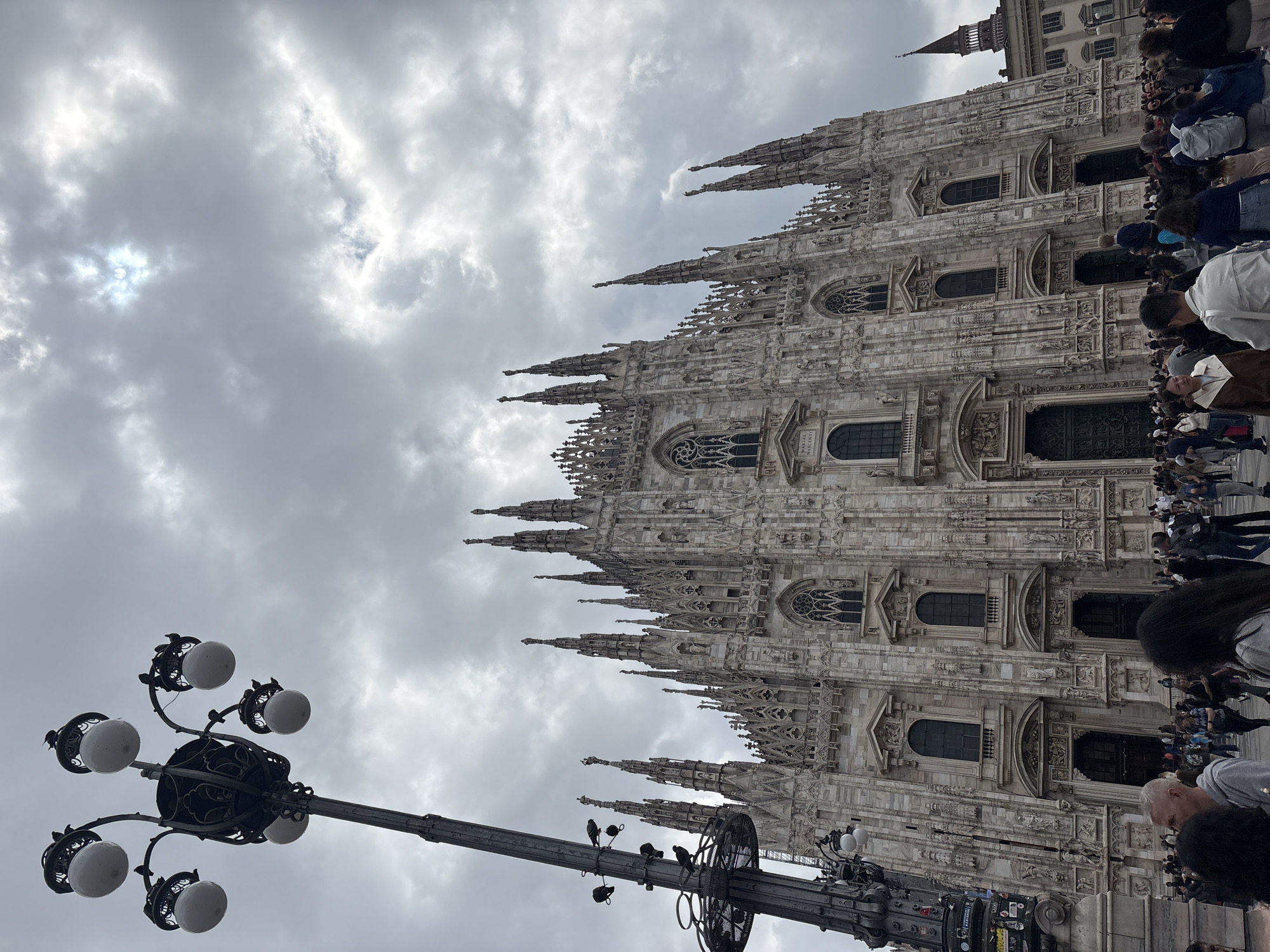 The image shows the Milan Cathedral in Milan, Italy, seen from a low-angle perspective. The cathedral's facade is ornate, with numerous spires and detailed carvings. A crowd of people is visible on the right side of the image, suggesting the cathedral is a popular tourist destination. A street lamp with multiple globe-shaped lights is positioned in the foreground, partially obscuring the view of the cathedral. The sky is overcast with grey clouds and the sun partially visible behind the clouds.