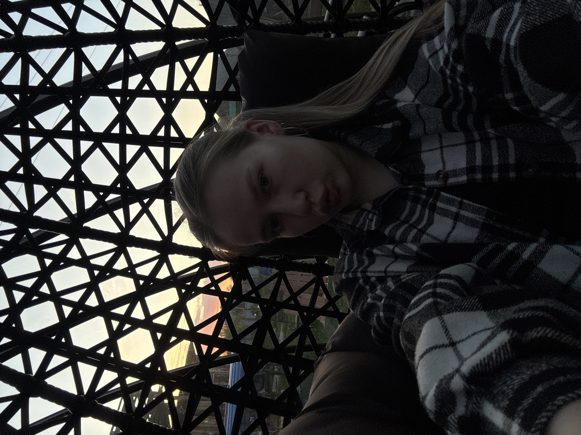A fair-skinned female is partially visible, lying in a dark-colored, intricately patterned, hanging chair. The chair's woven design creates a geometric grid against the backdrop of a muted sunset or sunrise. Through the chair's structure, a glimpse of a building and vegetation can be seen, potentially indicating an urban or suburban setting. The woman is wearing a dark and light-colored plaid shirt.
Although the photograph does not show the city in Zhigalki Russia, it has been included based on the request.