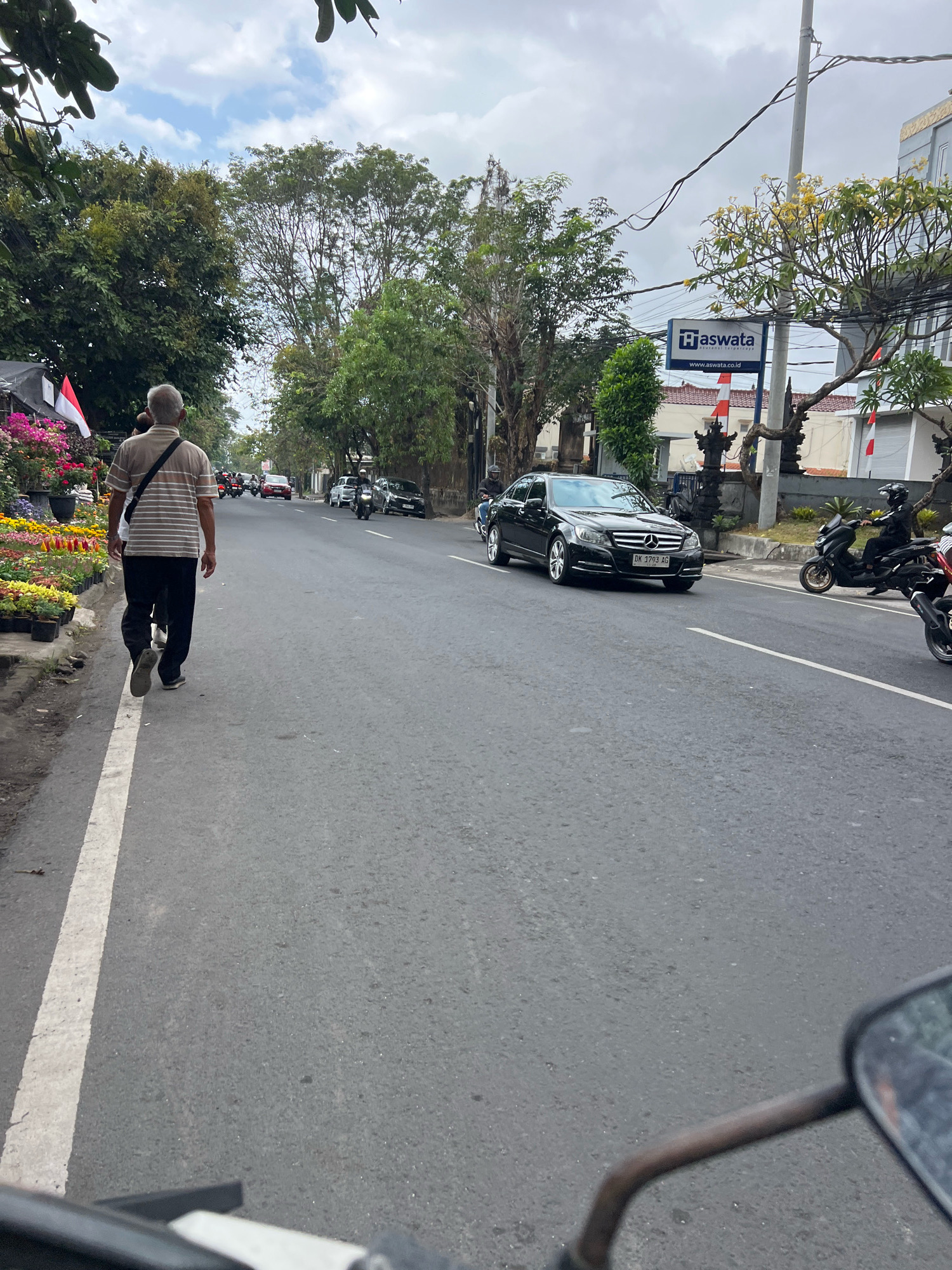 In Denpasar, Indonesia, on a cloudy day, a man walks along the side of a paved road. He is seen from behind, wearing a striped shirt and dark pants, with a bag slung across his shoulder. The road is lined with trees and buildings. A black Mercedes-Benz car is driving on the road, with its license plate visible. Several motorcycles are also present on the road. On the left side of the road, there are flower stalls with various colorful flowers. A sign for "Aswata" is visible on a building in the background.