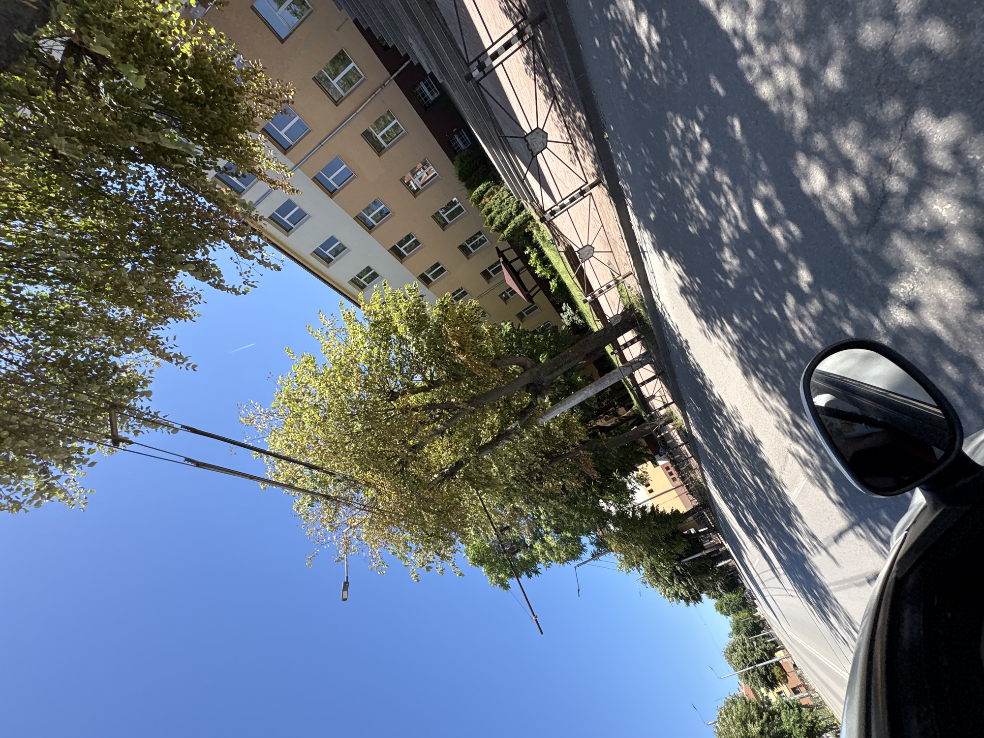 The image depicts a sunny day in Vratsa, Bulgaria, from the perspective of a moving vehicle. The scene is dominated by a light-colored, multi-story building with numerous windows, partially obscured by lush green trees. A road stretches into the distance, lined with more trees and a sidewalk with a decorative metal fence. Overhead, power lines and streetlights are visible against a clear blue sky. Shadows of trees are cast across the asphalt road, suggesting the sun is high in the sky. The perspective from the car includes a portion of the vehicle's side mirror and the edge of its bodywork in the foreground.