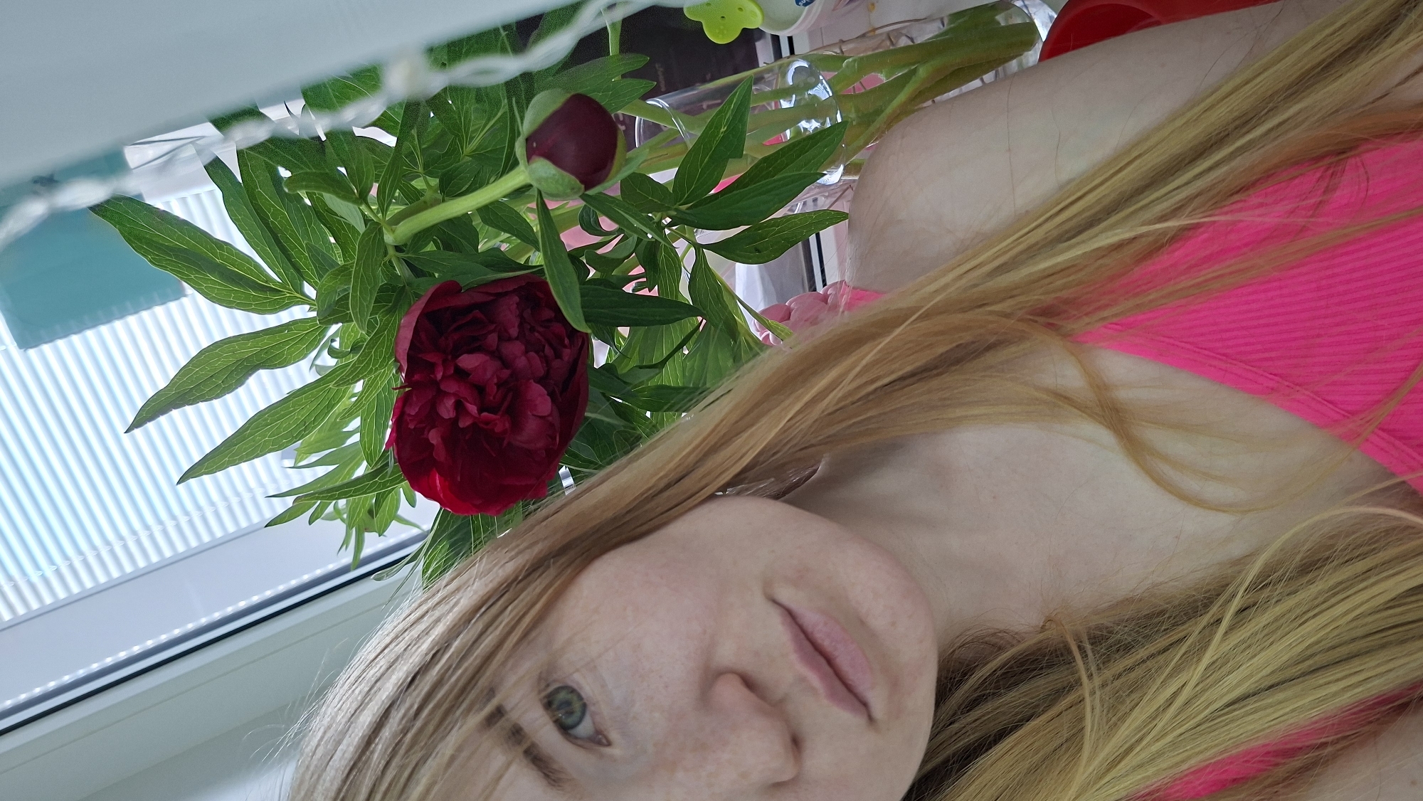 The image shows a woman with freckled skin, long blonde hair, and green eyes. She is wearing a bright pink top. A dark red peony flower and its foliage are positioned near her head. In the background, there is a window with white blinds and a blurry assortment of items. The location is Chaikovsky, Russia.