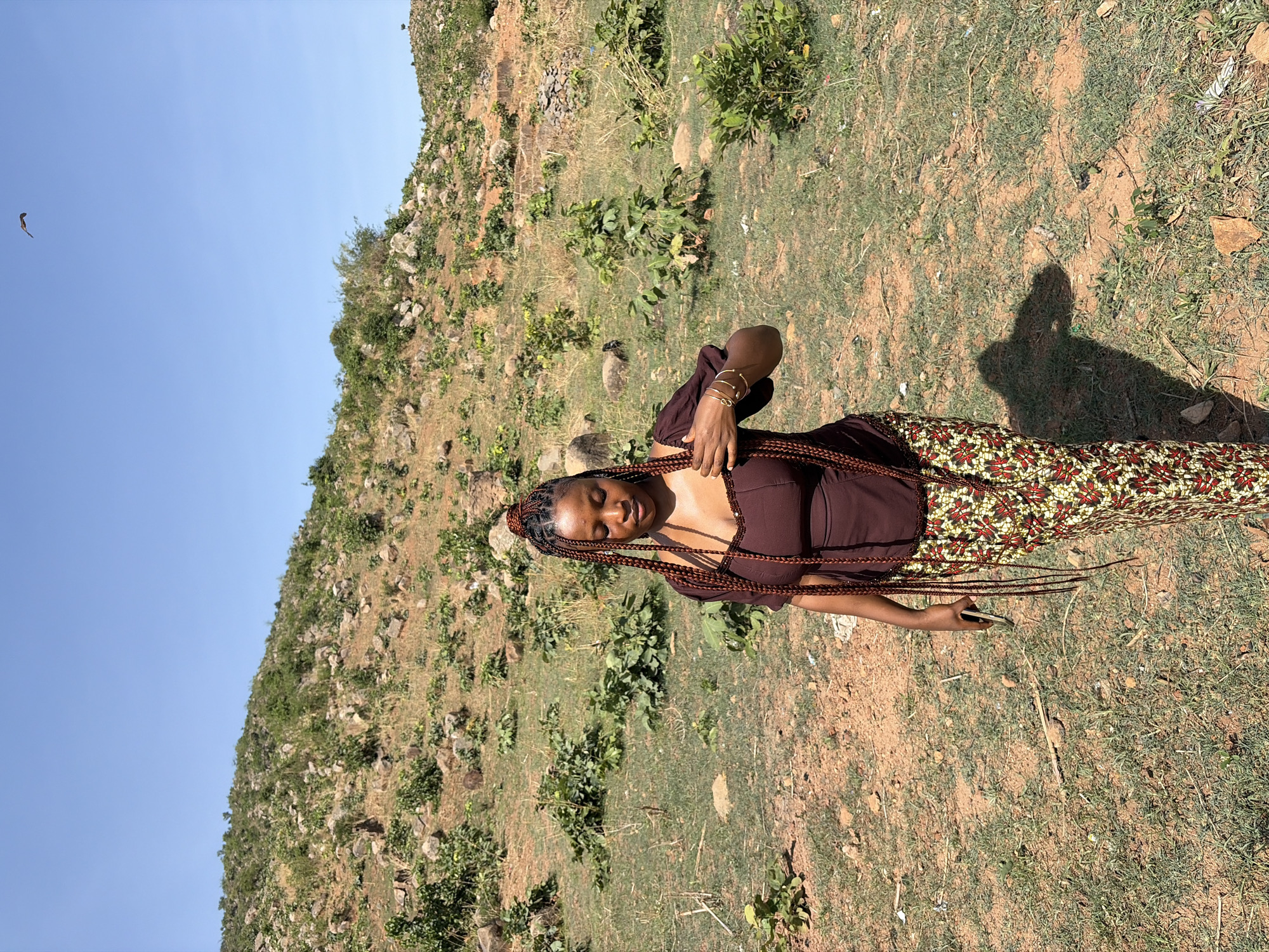 A young woman stands on a grassy, rocky hillside under a clear blue sky. She is wearing a brown top and a colorful patterned skirt. The hillside behind her is covered with rocks, shrubs, and grass. In the far left of the frame, there is a soaring bird. The photograph appears to have been taken outdoors in daylight. Based on the hillside vegetation, the location may be in Jos, Nigeria. The woman is holding a phone in her right hand.