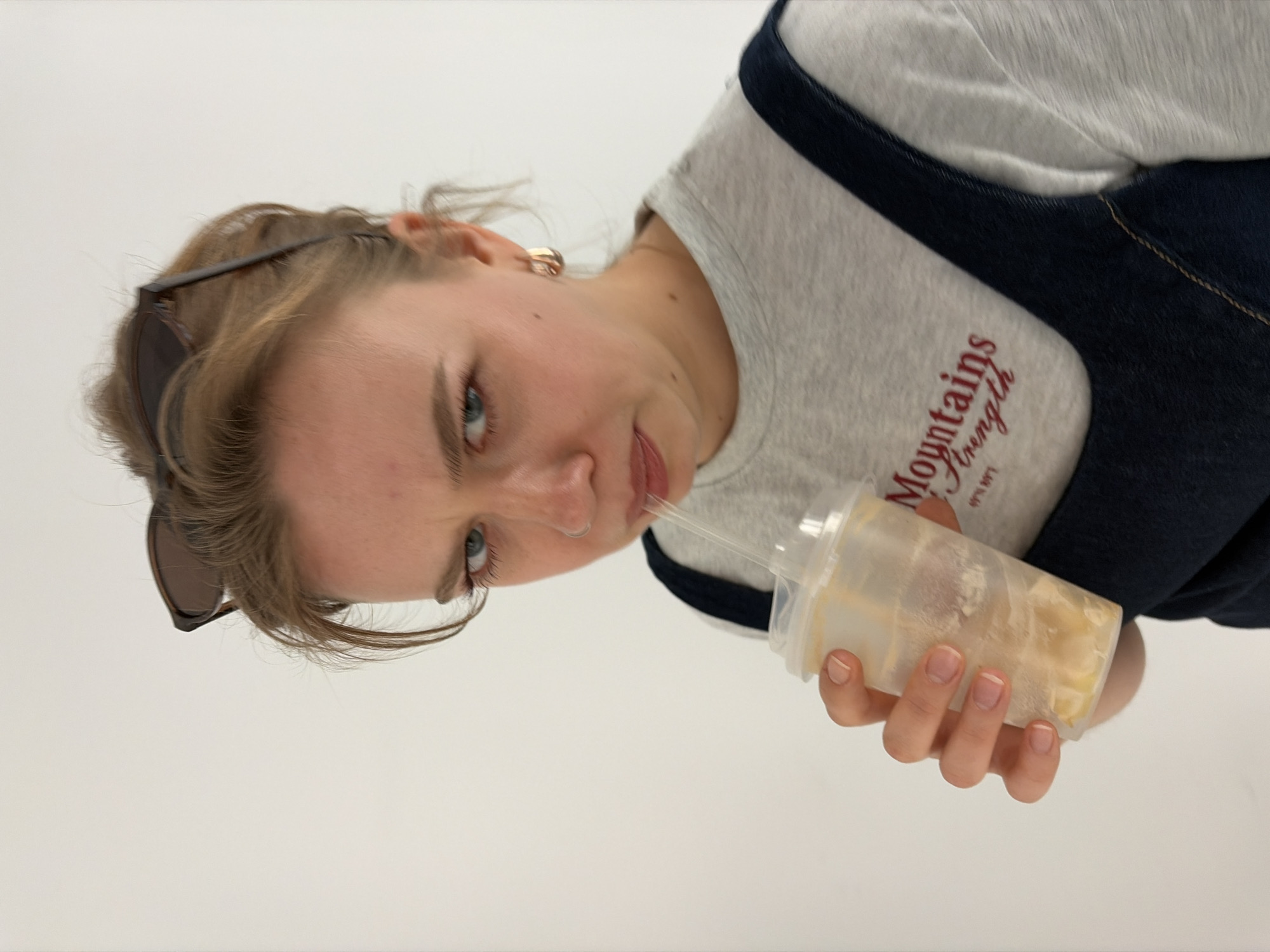 A young woman is drinking from a translucent plastic cup with a straw. She is wearing a gray shirt with the text "Mountains Strength" and a dark blue overall dress. Sunglasses are perched on her head. The background is plain white. The location is Бараўлянскі сельскі Савет, Belarus.
