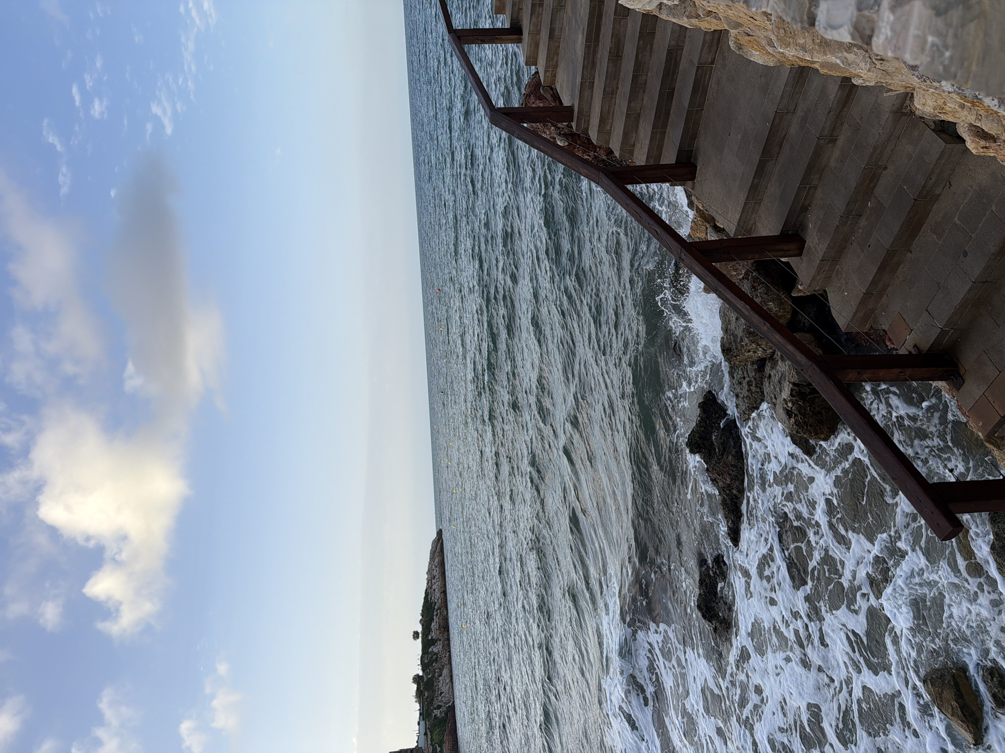 The image depicts a seaside scene in Salou, Spain. In the foreground, a stone staircase with wooden railings leads down towards the water. The sea is visible, with waves gently lapping against the rocky shore. In the background, a distant peninsula with some vegetation and structures can be seen, along with a clear blue sky dotted with scattered white clouds. The overall atmosphere is calm and serene.