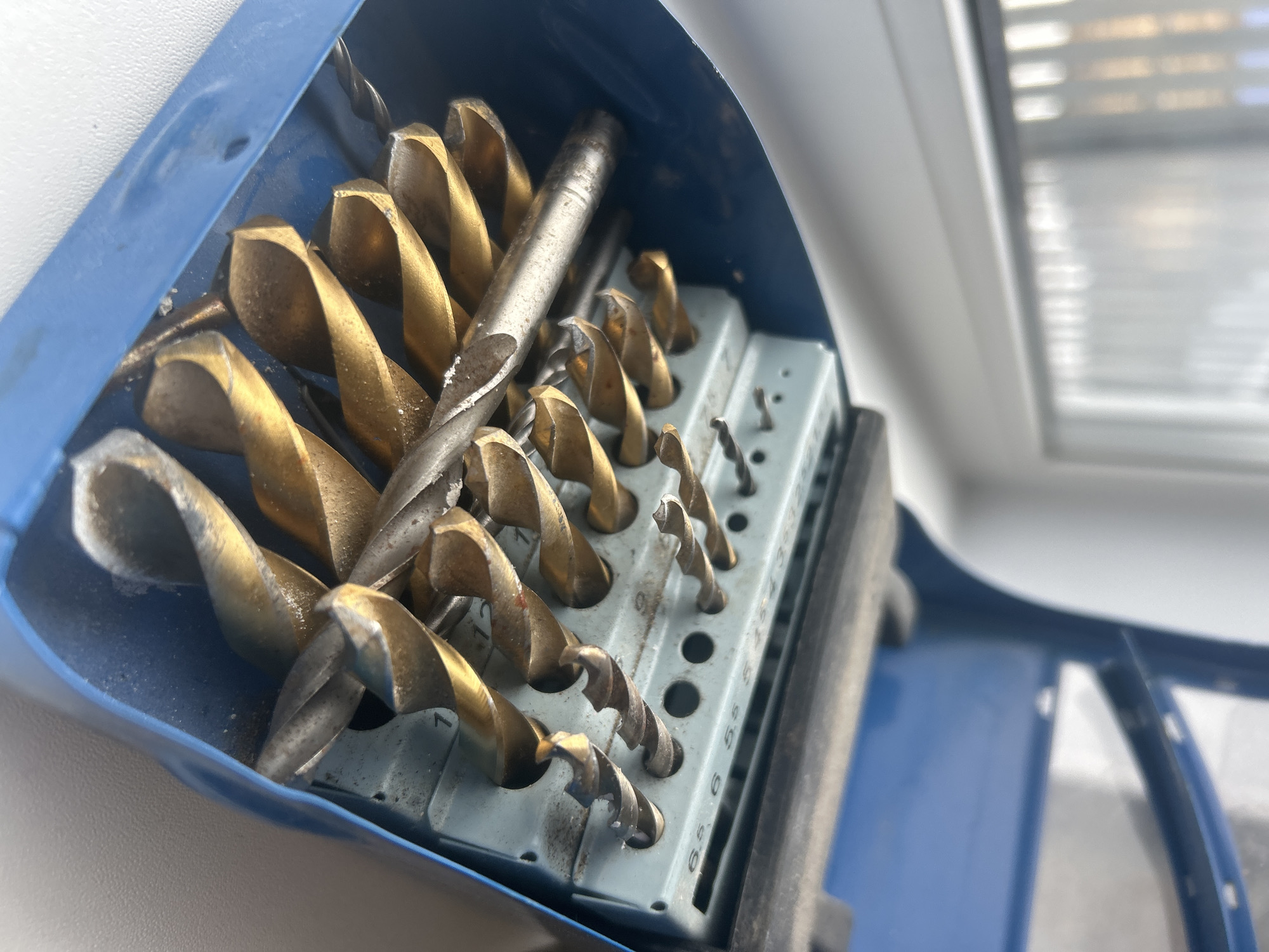A close-up shot depicts an open blue drill bit case containing an assortment of drill bits. The drill bits are primarily gold-colored, with one silver-colored bit placed amongst them. They are arranged within a gray holder with labeled sizes. The setting appears to be indoors, possibly near a window, as suggested by the blurred background showing a window with blinds. There is no indication of the location being in Chaikovsky, Russia. There are no people visible.