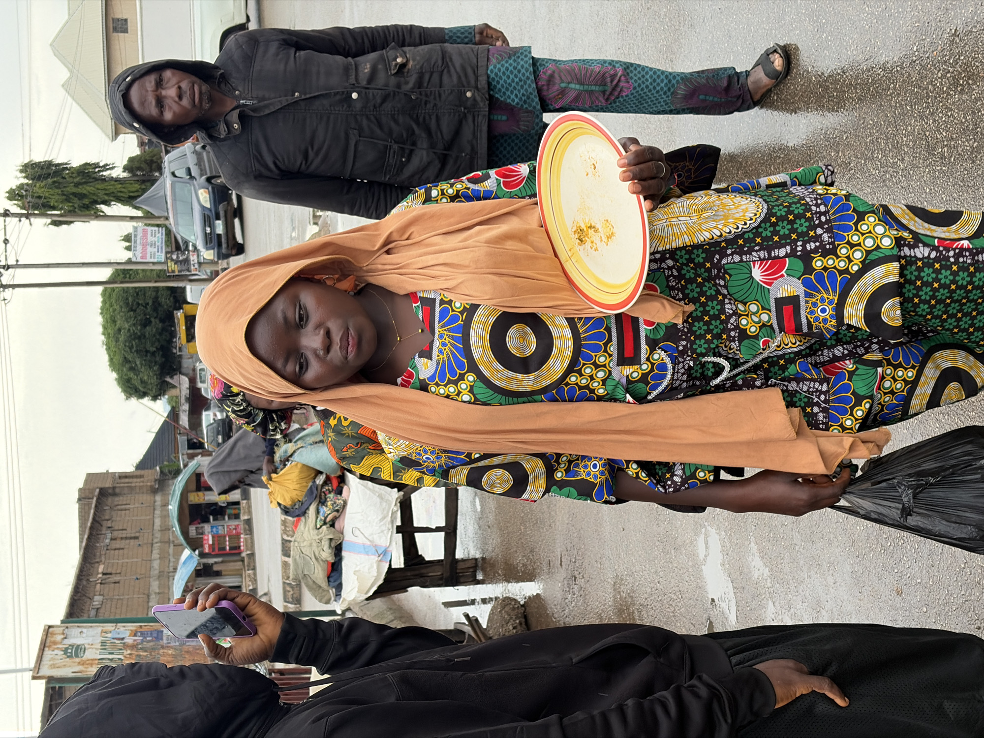 The image depicts a street scene in Jos, Nigeria. In the foreground, a person in a black hooded jacket is holding a purple smartphone. Behind them, a young woman wearing a colorful, patterned dress and a peach headscarf is holding a plate. Her expression is somewhat serious, and she is looking directly at the camera. To her right, a man wearing a black jacket with a hood is visible, with his hands in his pockets. He has a somber expression.
The background shows a street with a wet, reflective surface, suggesting recent rain or damp conditions. There are buildings and market stalls, with some clothing and other goods displayed. A dark-colored car is parked on the street. The overall lighting suggests it is daytime, possibly overcast or after rain, as the sky is bright but not intensely sunny. The presence of market stalls and the style of dress indicate a local Nigerian setting. The interaction between the individuals, particularly the young woman holding the plate and looking at the camera, might suggest a moment captured during a transaction or interaction in the market. The wet ground and overcast sky contribute to a somewhat muted atmosphere.