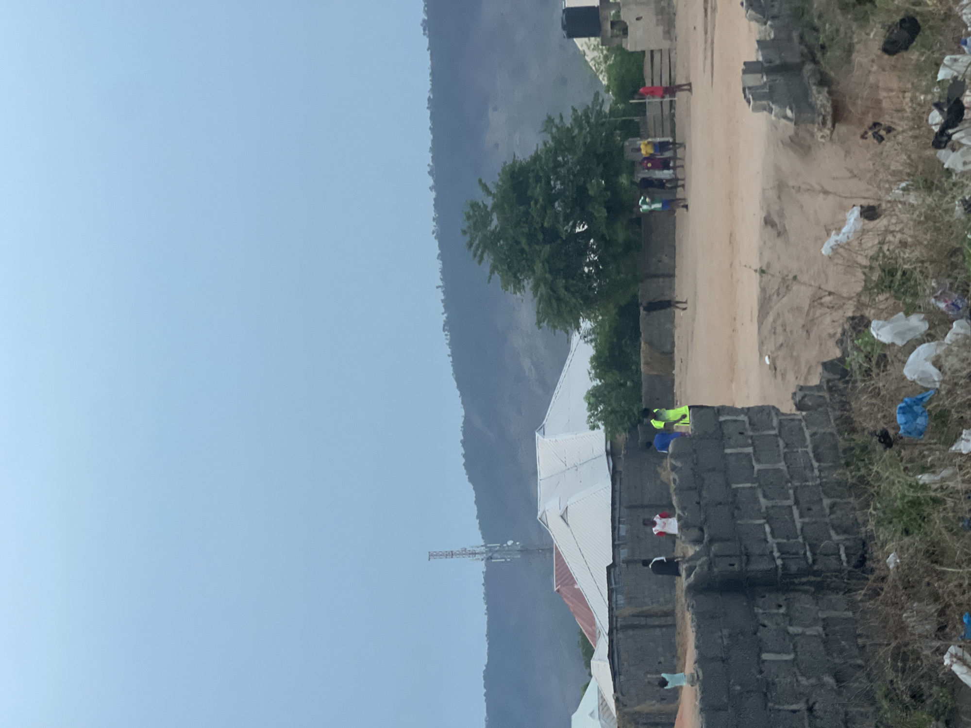 The image depicts an outdoor scene in what appears to be a semi-urban area of Lokoja, Nigeria, under a light, possibly overcast sky. In the background, a large range of hazy, dark mountains extends across the horizon. A tall, slender communication tower stands prominently on the left, silhouetted against the distant hills.
In the mid-ground, several simple buildings with corrugated white and reddish-brown roofs and grey concrete block walls are visible. A significant green tree is situated near these structures. A dirt path cuts through the right side of the scene, where a group of individuals is gathered, appearing to socialize informally. They are dressed in casual, brightly colored attire, including red, yellow, and blue shirts.
The foreground features a low wall constructed from grey concrete blocks on the right. The ground in this area is dry and dusty, noticeably strewn with a considerable amount of plastic bags and other refuse, indicating a challenge with waste management. More people are visible closer to the foreground, one in a red and white shirt, another in black, and a third in a light blue shirt. A black water tank is visible on top of a building in the upper right.
The overall atmosphere suggests a casual community gathering. The diffused lighting implies it could be early morning, late afternoon, or an overcast day. No discernible text is visible in the image.
