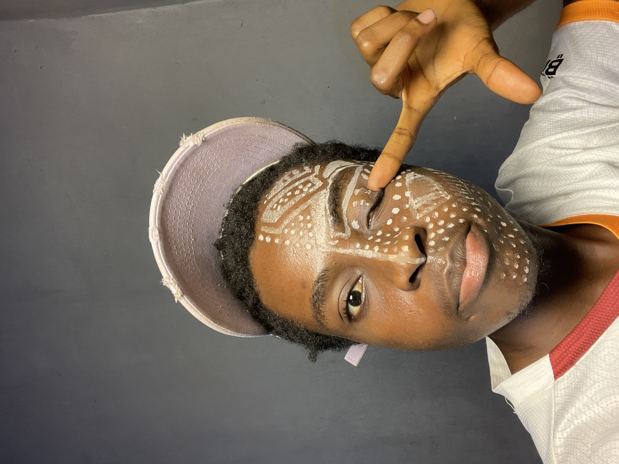 A young Black man with dark hair, wearing a light purple hat and a white and orange sports jersey, has white paint markings on his face. He is positioned against a dark gray wall. He is looking at the camera, with his right index finger pointing at his face above his right eye. The man's face features geometric patterns and dots painted in white. The photo appears to be taken indoors, with the background consisting of a plain, dark gray wall. There is no direct indication that the image was taken in Nkpor, Nigeria from the image alone.