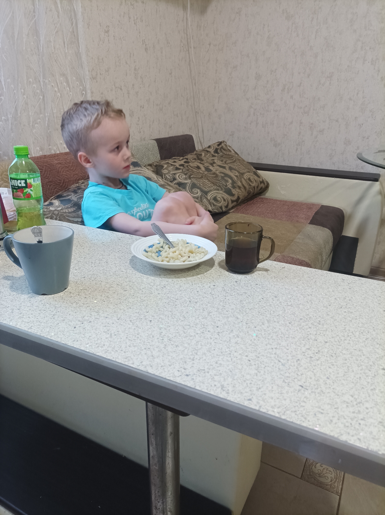 A young boy sits on a couch in a room, seemingly in Ilskiy, Russia, as indicated by the Cyrillic text on the drink bottle. He wears a blue t-shirt and has blonde hair. The boy appears to be deep in thought, with his arms wrapped around his legs. On the table in front of him are a blue mug with a spoon inside, a plate of pasta with a spoon, and a brown mug. A bottle of juice sits to the left of the mug. The couch has multiple pillows with different patterns and colors. The wallpaper in the room is a light color with a subtle pattern. A metal table leg is visible supporting the white tabletop.