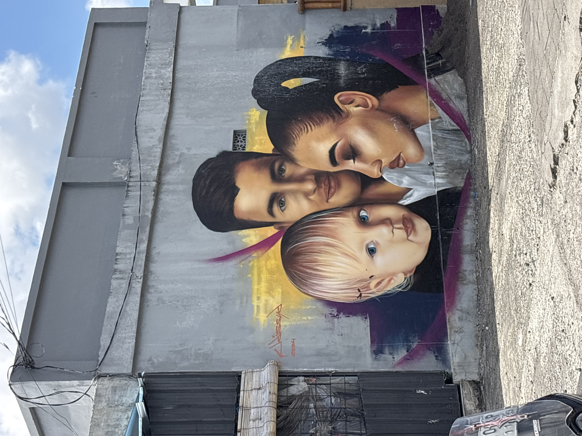 A mural depicting a family of three graces a gray, multi-level building in North Kuta, Indonesia. The mural features a woman with dark hair tied back, a man with short dark hair, and a light-haired child with blue eyes. They are clustered close together in a loving embrace. The mural is painted with a combination of realistic and abstract techniques, featuring blended colors and painterly strokes. The artist's signature "Kalisya" and the year "2024" are visible near the bottom of the mural. Below the mural, there's a metal rolling shutter, and glimpses of the street are seen. Electrical wires are visible on the upper left of the building. The sky above is blue with scattered clouds. The overall setting suggests an urban environment.