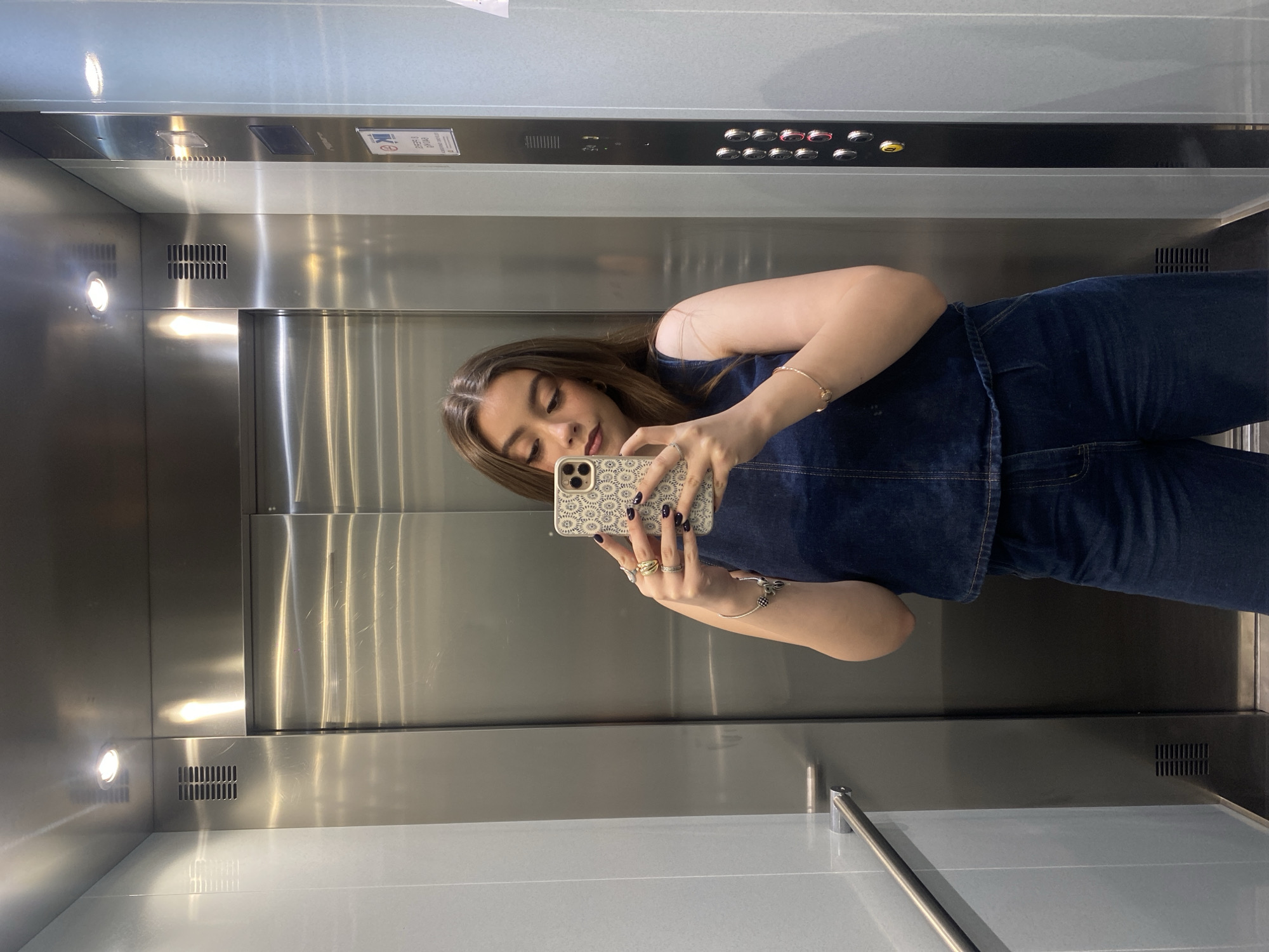 A young woman is taking a selfie in what appears to be the elevator of an Unknown building in Unknown. She is wearing a dark blue denim top and jeans, and several rings and bracelets are visible on her left hand. The elevator has a stainless steel interior, push-button controls, and a horizontal handrail.