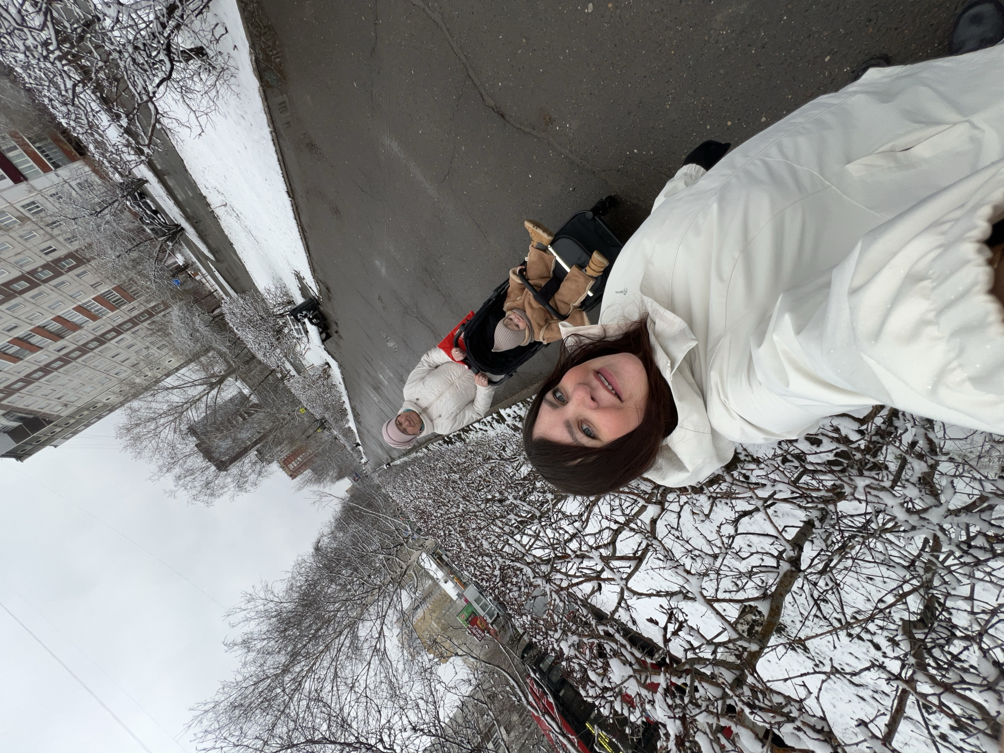 Here's a detailed description of the image:
**Scene:** An outdoor selfie taken on an overcast day in Perm, Russia. The ground is covered in snow, particularly on the trees and bushes, suggesting a winter setting. A wet pavement road is visible.
**People:** Three people are in the image.
* A woman with brown hair is holding the camera for a selfie. She wears a white jacket.
* An older woman is pushing a stroller. She also wears a white jacket and a pink hat.
* A baby sits in the stroller, bundled up in a beige outfit.
**Objects:**
* Stroller
* Buildings, including a multi-story apartment building with red accents.
* Trees and bushes covered in snow
* Wet road
**Actions & Interactions:**
* The woman is taking a selfie.
* The older woman is pushing the stroller.
**Environment:**
* The scene is in an urban area, likely a residential neighborhood in Perm.
* The weather is overcast, with snow or ice present.