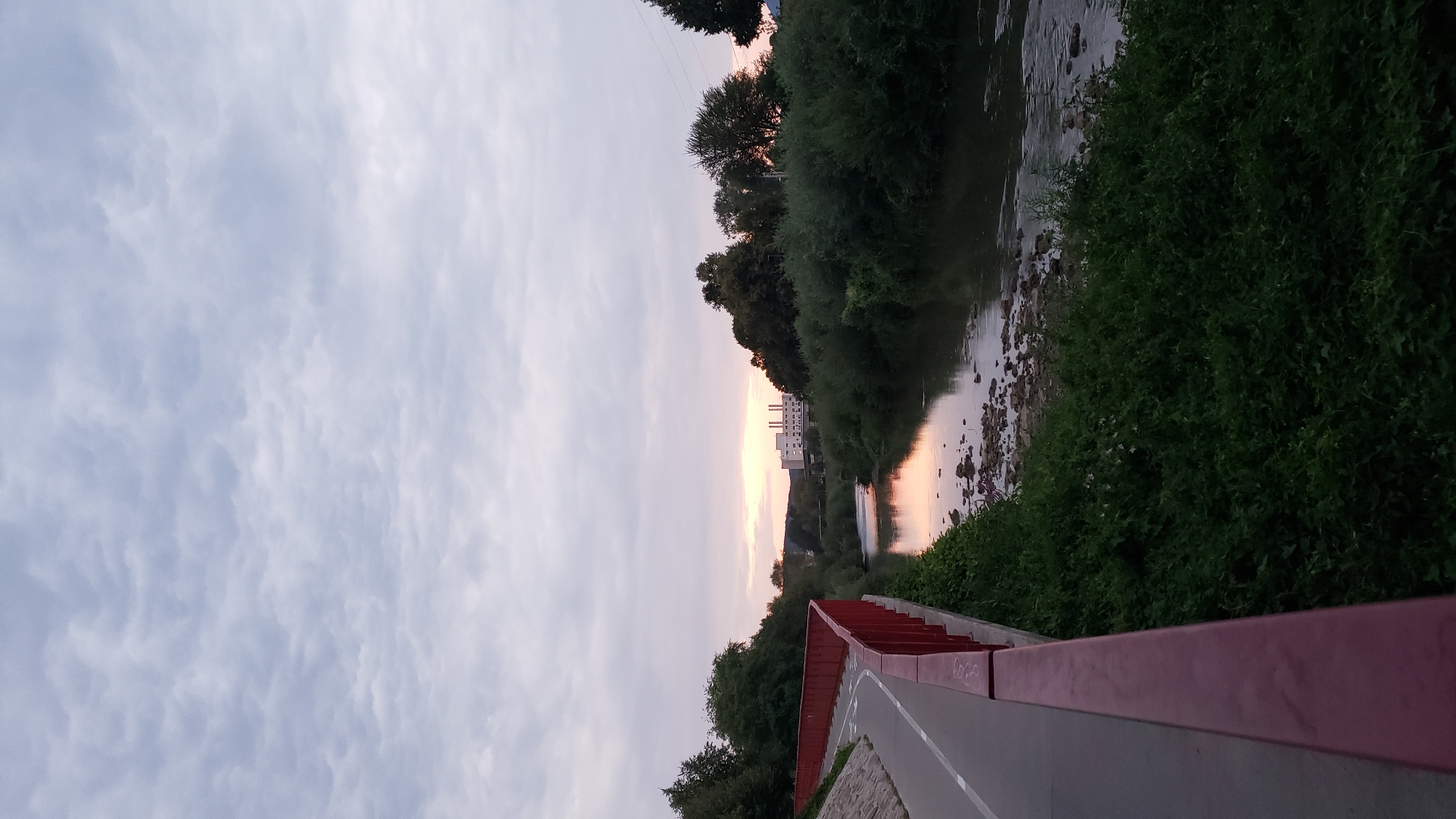 The image depicts a pathway bordered by a red railing on the left, curving away from the viewer. The pathway is paved and has white lines indicating lanes. Beyond the railing, dense green foliage covers a slope leading down to a slow-moving river. The river reflects the soft colors of the twilight sky, with hues of pink and orange visible. Across the river, a line of trees and bushes forms a natural barrier, with a building complex featuring multiple white structures and smokestacks visible in the distance, situated on a slight rise. The sky above is filled with a dense layer of grey, textured clouds, indicative of overcast weather or the approach of evening. The scene is serene and quiet, with no people or animals present. The location is likely a park or recreational area along a river in Prešov, Slovakia, given the architectural style of the building complex and the general landscape, which is consistent with urban environments in Central Europe.