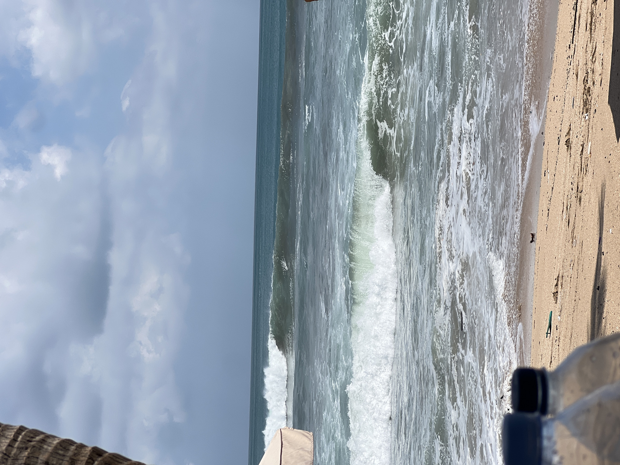 The image shows a beach in Kuta, Indonesia, with waves crashing on the shore. The sky is cloudy, and the ocean is a deep blue-green color. In the foreground, there are two plastic water bottles on the sand. There are also some pieces of debris scattered on the beach. A large palm tree trunk is visible on the left side of the image. A tan-colored umbrella or canopy is partially visible on the left. The scene appears to be a natural beach setting with some man-made elements present.