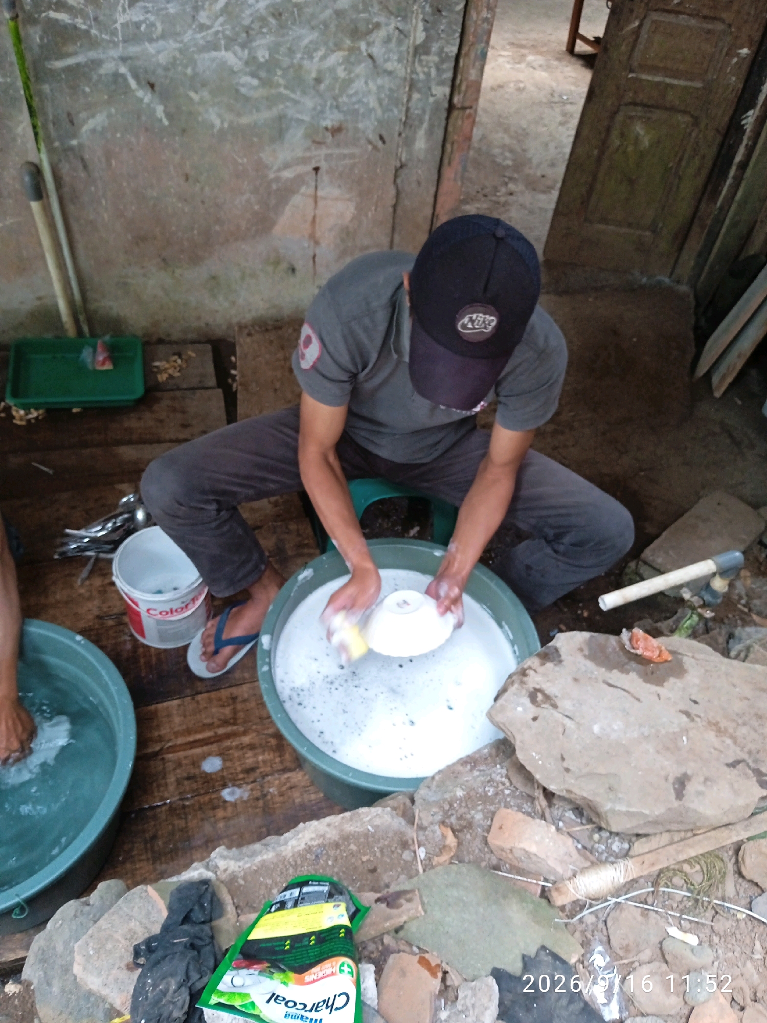 A person wearing a dark t-shirt and pants, and a dark baseball cap, is washing dishes in a large green tub filled with soapy water. The person is seated on a small green stool. To the left of the tub is a white bucket with a red label. In the background, there is a weathered wall, a wooden door, and some stacked wood. In the foreground, there are large rocks and debris, and a green package of what appears to be charcoal briquettes. The scene appears to be outdoors.