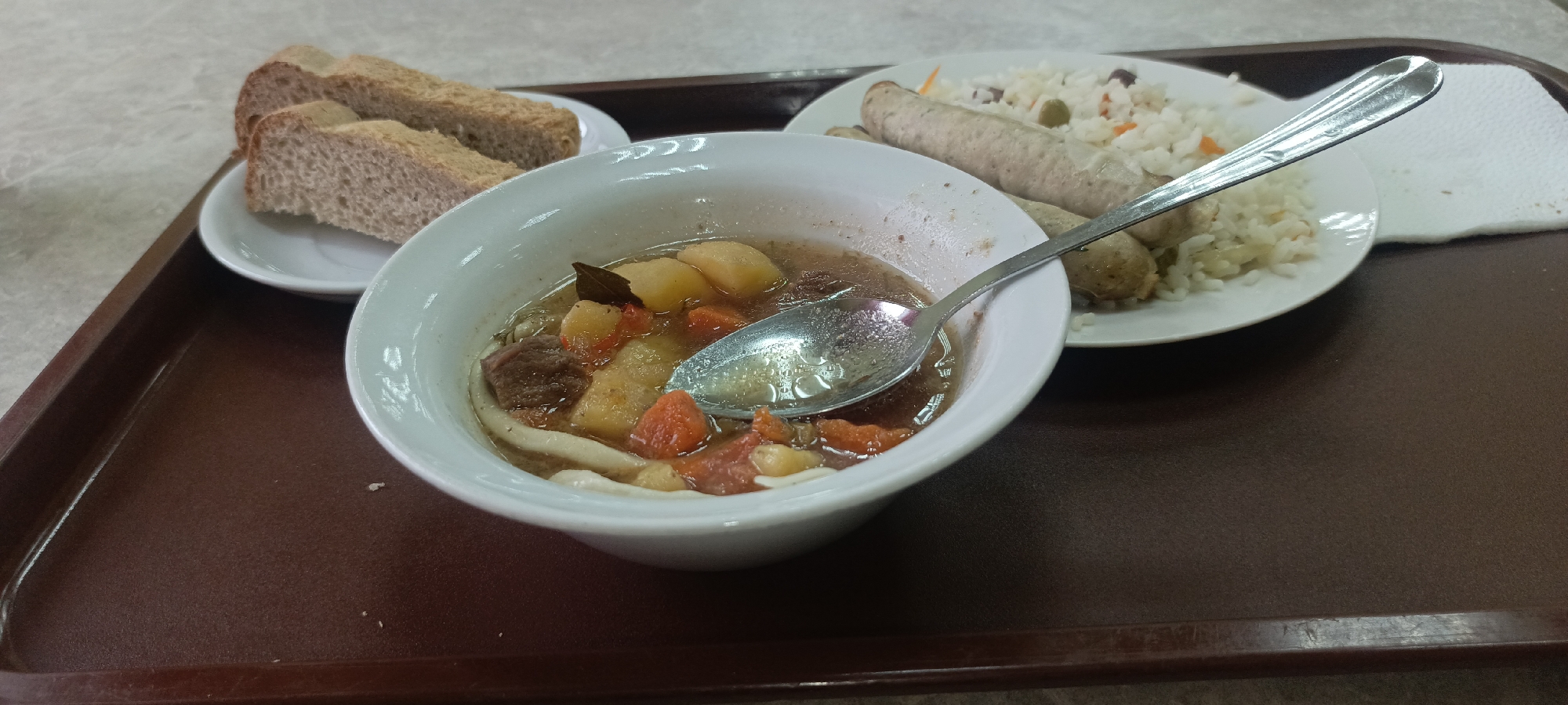 The image depicts a meal on a brown plastic tray. There is a white bowl of soup with beef, potatoes, carrots, and noodles, with a metal spoon resting inside. Adjacent to the soup is a small plate of brown bread slices. Another white plate holds sausages and rice with vegetables, next to a folded white napkin. The tray sits on a beige patterned surface. No people are visible in the image.