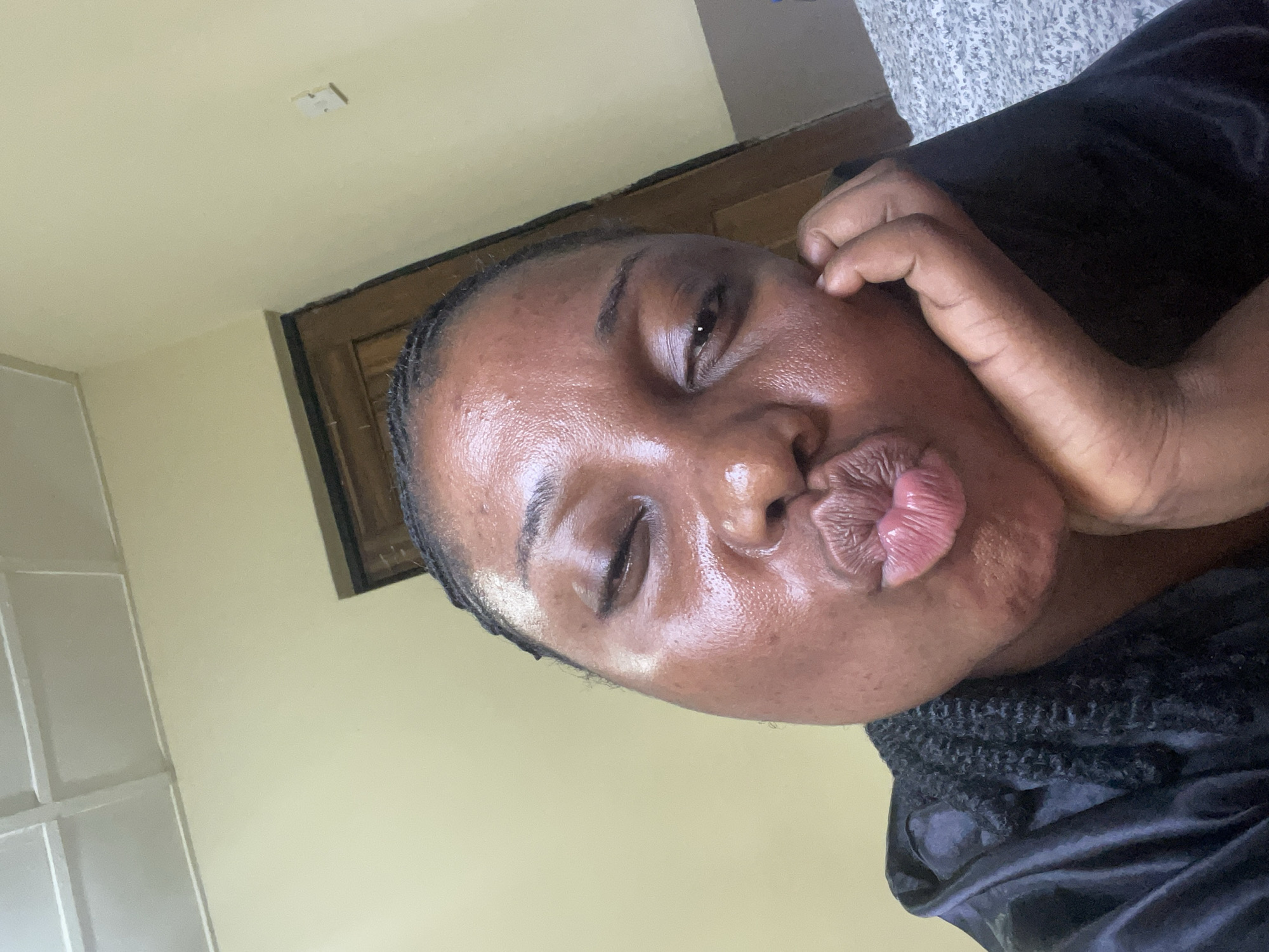 A person is close to the camera, with their head tilted to the side, and their lips puckered as if kissing. The background is blurry and shows parts of a wall, a door, and what looks like a window. The image does not provide adequate context to infer a precise location in Jos, Nigeria.