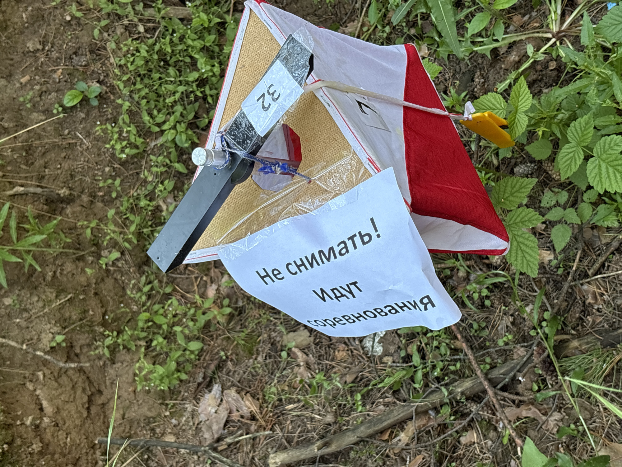 A medium shot captures a control point marker, commonly used in orienteering, situated in a natural outdoor environment. The marker itself is a red and white triangular prism with a black and silver top structure. A sign, written in Russian, is attached to the marker, stating "Не снимать! Идут соревнования," which translates to "Do not remove! Competition in progress." A yellow tag hangs from the marker. The ground around the marker is covered in dirt, dried leaves, pine needles, and various green plants and grasses. The background consists of more vegetation and soil, suggesting a wooded or park-like area. There is no indication of the city of Chaikovsky, Russia, within the image itself.