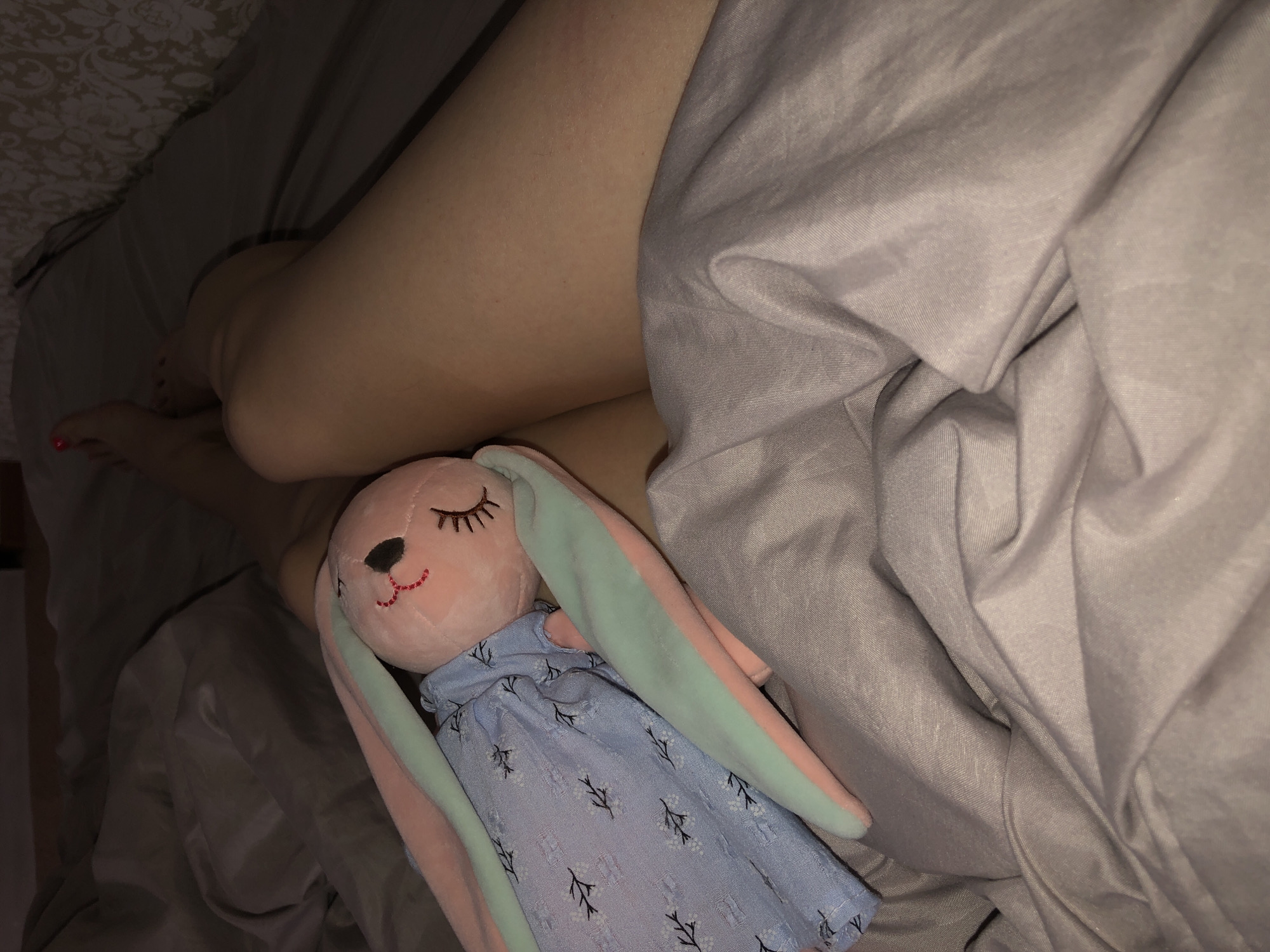 A person's bare legs are visible under a gray comforter, with a pink plush rabbit wearing a blue dress positioned on the bed. The setting appears to be a dimly lit bedroom. The location of Noviy, Russia is not relevant as the context of the image does not provide that information.