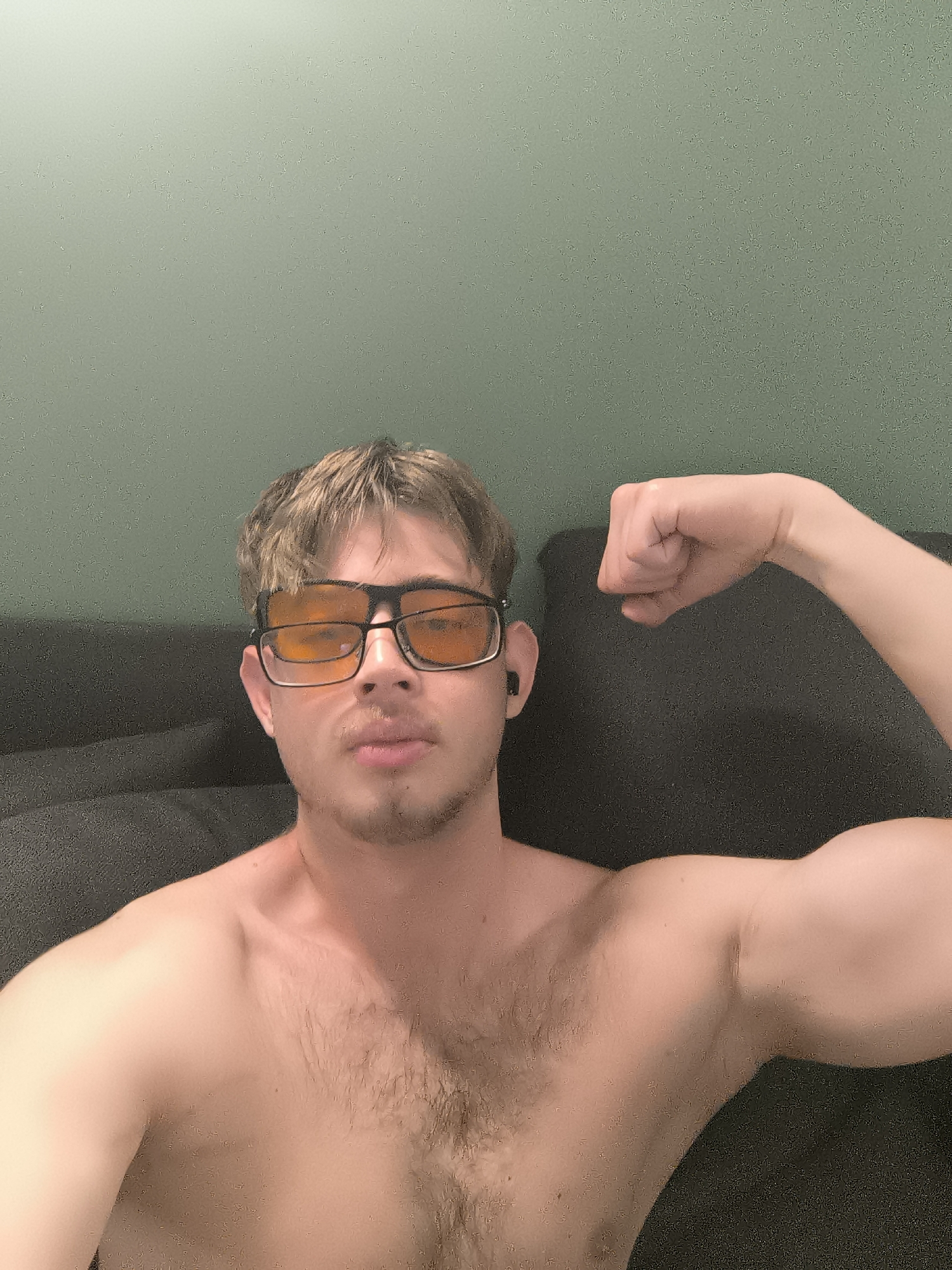 A young man is depicted in a medium shot, flexing his left bicep. He is shirtless, revealing a lightly haired chest. He has light brown hair, a faint mustache, and a goatee. He is wearing prominent black-framed glasses with amber or orange-tinted lenses, and a black earbud is visible in his right ear. His expression is neutral, looking directly at the camera.
The scene appears to be indoors, possibly in a living room, in Sofia, Bulgaria. The background features a plain, light green wall at the top, transitioning into a dark, textured surface, likely a sofa or couch, behind the man. The lighting suggests an indoor setting, possibly with artificial light, given the warm hue from the glasses. There is no visible text in the image.