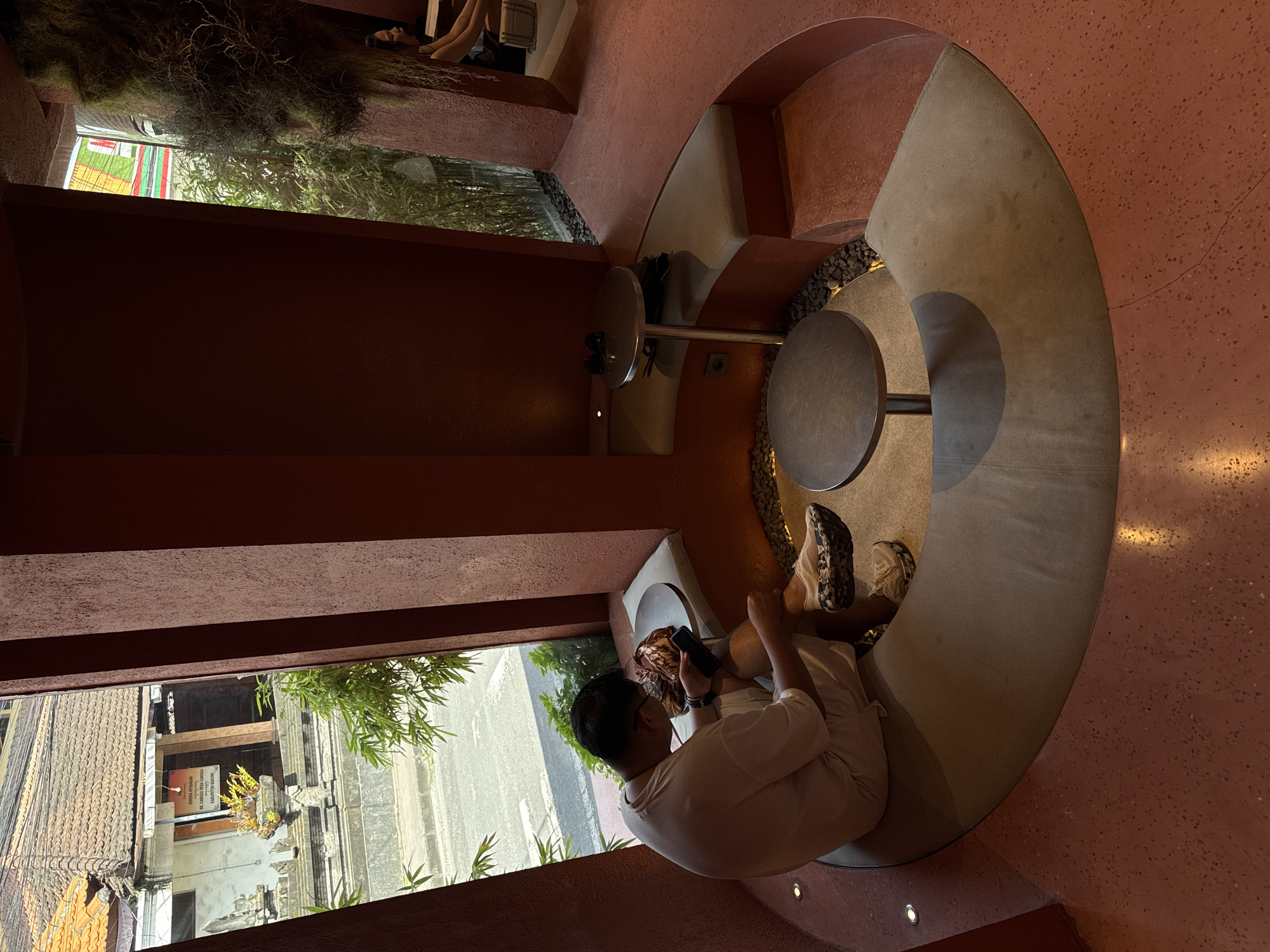 A person is sitting in a curved booth reading a cellphone, inside a building with a window view in North Kuta, Indonesia. The person is dressed in light colored casual clothing. They are seated at a round table within the booth. The building has red-brown colored walls and flooring with interspersed small pebbles. The booth is built-in and semi-circular. Outside the window is a street view. There is hanging foliage near the ceiling.