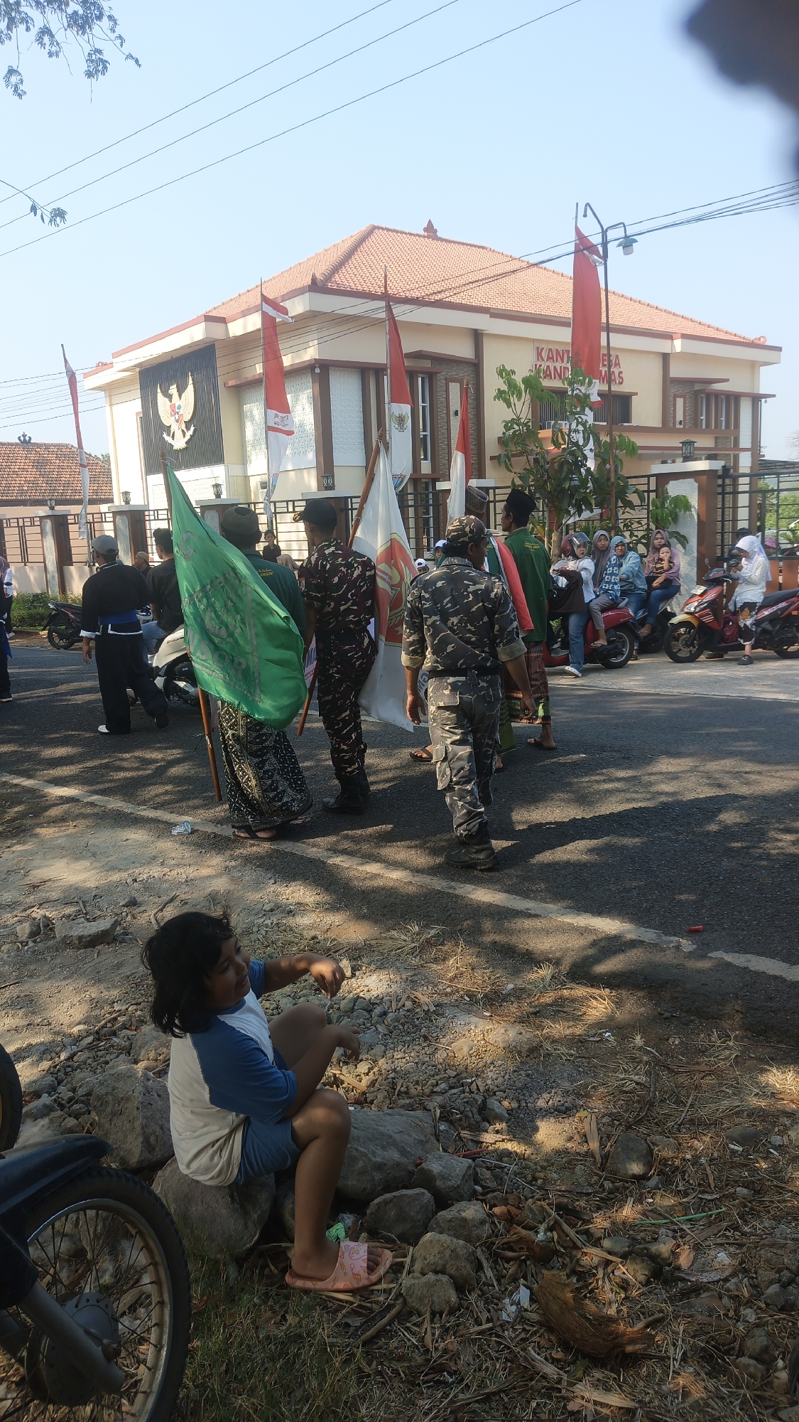 The image depicts an outdoor scene in Kandangmas, Indonesia, featuring a building identified by signage as "KANTOR DESA KANDANG MAS". Several individuals are present, some holding flags, including Indonesian flags (red and white) and a green flag. The individuals are dressed in various attire, including military-style camouflage uniforms, traditional Indonesian clothing, and casual wear. Some are standing, while others appear to be walking. In the foreground, a young child sits on rocks next to a motorcycle. The environment includes a paved road, dry grass, trees, and the building, which has an orange tiled roof and architectural details common in Indonesia. The sky is clear and blue, suggesting a sunny day. The overall atmosphere appears to be that of a gathering or event.