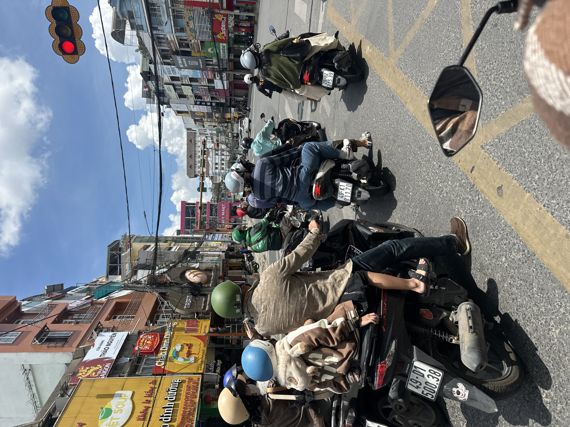 This is a medium shot, eye-level, daytime, outdoor, street-level, full-color photograph taken on a bright, cloudy day in Phường Xuân Hương, Đà Lạt, Vietnam. The scene depicts a busy intersection where multiple motorcycles are stopped at a red traffic light.
In the foreground, on the right, the handlebar and rearview mirror of a motorcycle are visible. The mirror reflects a checkered brown and white patterned blanket.
The central focus is a line of motorcycles and their riders at the intersection. On the left, a motorcycle is seen from the rear, with a person in a brown jacket and a child in a brown hoodie on the back. The child is wearing a blue helmet and the adult is wearing a tan helmet. The motorcycle has a license plate that reads "49-D1 500.38".
Next to them, a man wearing a green helmet and a patterned shirt is on his motorcycle. To his right, a person in a grey helmet and a blue and white striped shirt is also on a motorcycle. Further back, other individuals on motorcycles are also visible, some with green helmets.
The background shows a street lined with multi-story buildings, many of which have vibrant yellow and red signage. The buildings have a mix of architectural styles, and electrical wires are strung across the street. A traffic light displaying a red signal is prominently positioned in the upper right of the frame. The road surface is asphalt with yellow lane markings. The sky is a mix of blue and fluffy white clouds.