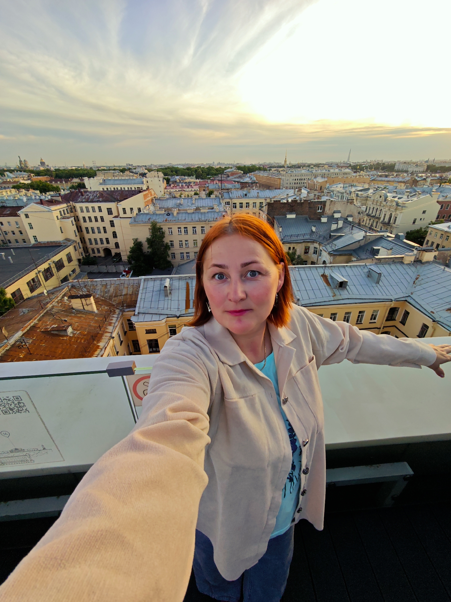 A medium shot captures a fair-skinned woman with auburn hair taking a selfie on a rooftop overlooking the city of Saint Petersburg, Russia. The woman is wearing a light-colored jacket over a blue t-shirt and dark pants. Her left arm is extended, holding the camera, while her right arm is bent at the elbow and resting on a railing. She is looking at the camera with a neutral expression.
The background reveals a dense urban landscape with numerous buildings of varying architectural styles, typical of Saint Petersburg. The rooftops are a mix of grey, red, and brown, with some featuring visible chimneys and vents. In the distance, several prominent landmarks, including domes and spires, punctuate the skyline, indicative of the city's historical significance. The sky above is partly cloudy with streaks of sunlight, suggesting it is either sunrise or sunset.
In the foreground, a portion of a railing and a informational display with a QR code are visible. The informational display appears to be mounted on a white surface. The overall scene suggests the woman is at a viewpoint or observation deck, enjoying a panoramic view of the city.