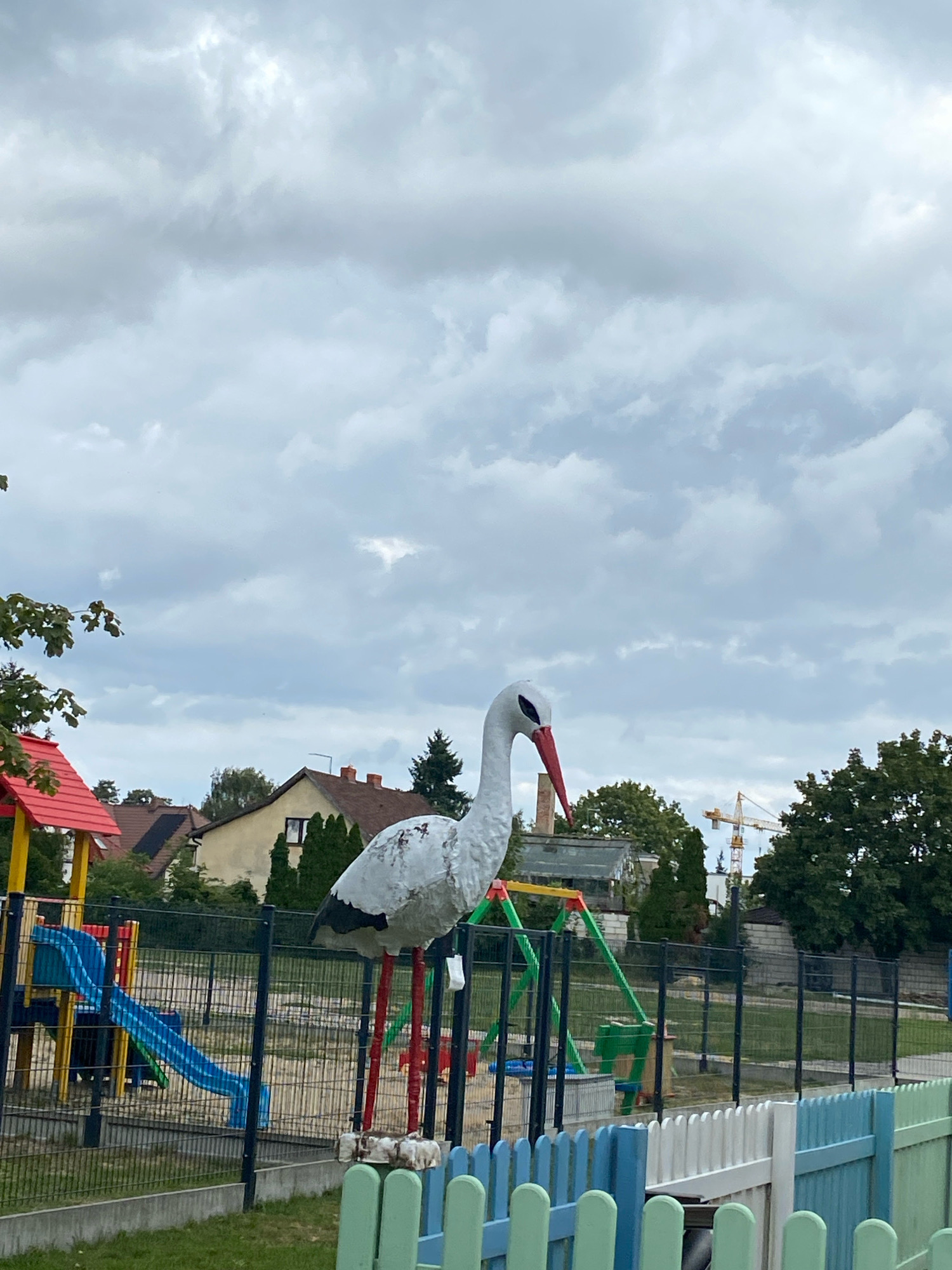 An outdoor shot shows a playground with a large, white stork statue as the central figure. The stork has a red beak and black markings on its wings and tail. It stands on red legs, one of which is supported by a wooden block. Behind the statue, a blue and green slide structure is visible, along with a swing set. Houses with tiled roofs and trees are in the background, under a cloudy sky. A yellow construction crane can be seen in the distance. The playground is enclosed by a black metal fence, and a colorful picket fence runs along the foreground. The image is taken in the city of Buk, Poland. There are no people visible in the image.