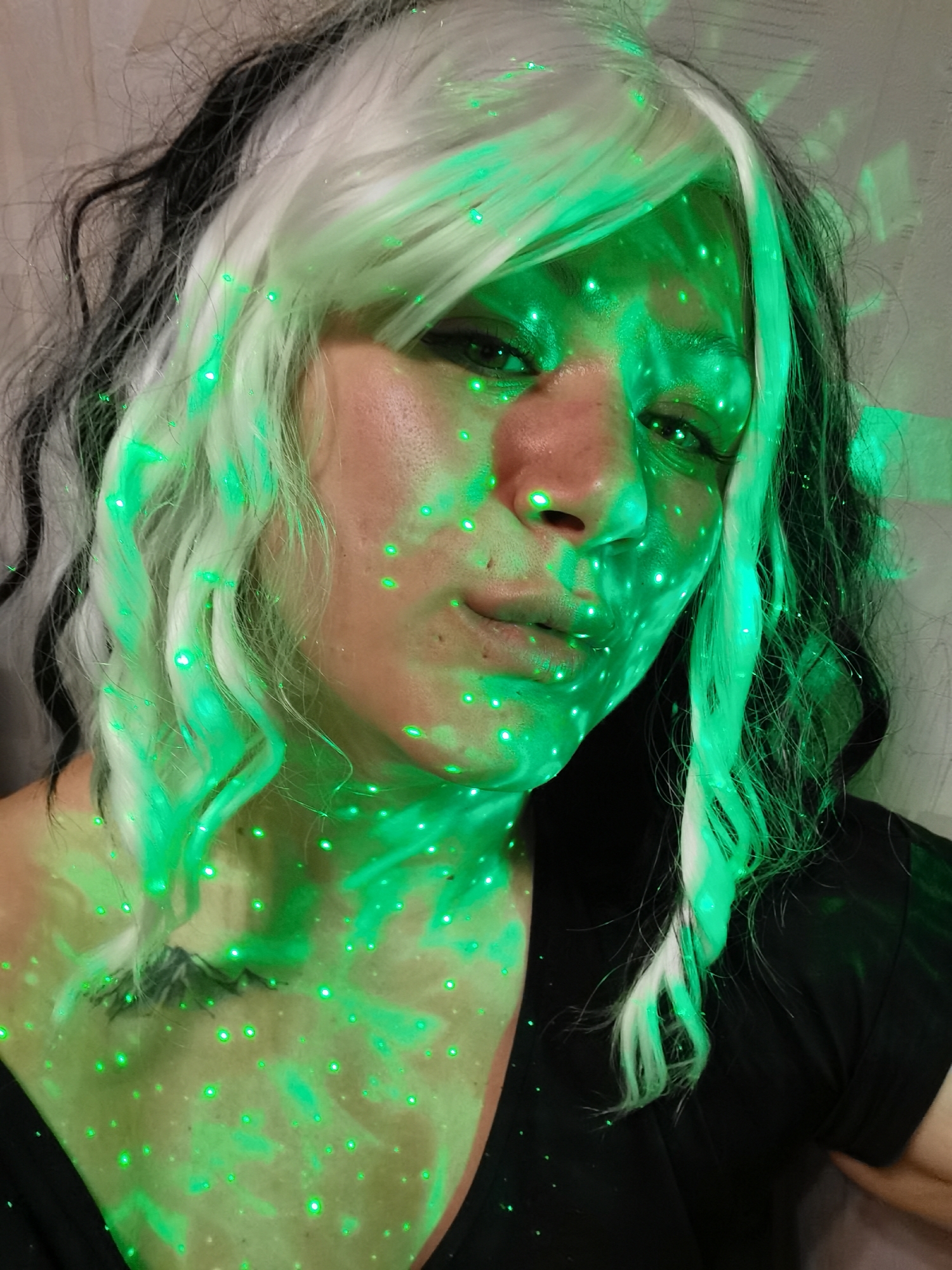 A close-up studio shot depicts a woman with fair skin illuminated by a green laser light. Her hair is a wig style, featuring a mix of white and black strands with some curls. She is wearing a black V-neck shirt. The green laser light creates a pattern of dots on her face, hair, and chest. No location information is discernible in the image.
