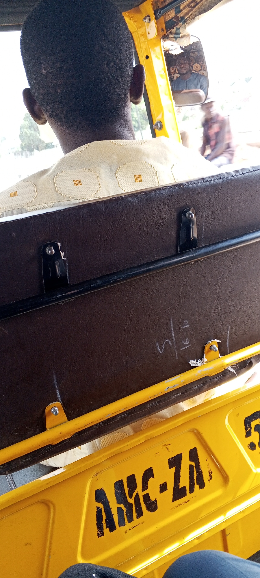 The photo shows the interior of a yellow and brown auto rickshaw, possibly a Bajaj RE, in Uke, Nigeria. The back of a dark-skinned person's head is in the foreground, wearing a light-yellow patterned shirt. The driver is visible in the rearview mirror. The back of the seat is brown and has a metal bar across it, with markings that appear to read "4/16" or similar handwritten numbers. The yellow body of the rickshaw has the black stenciled letters "AMC-ZA" visible. Another person is slightly out-of-focus through the rear of the vehicle. The general environment appears bright and outdoors.