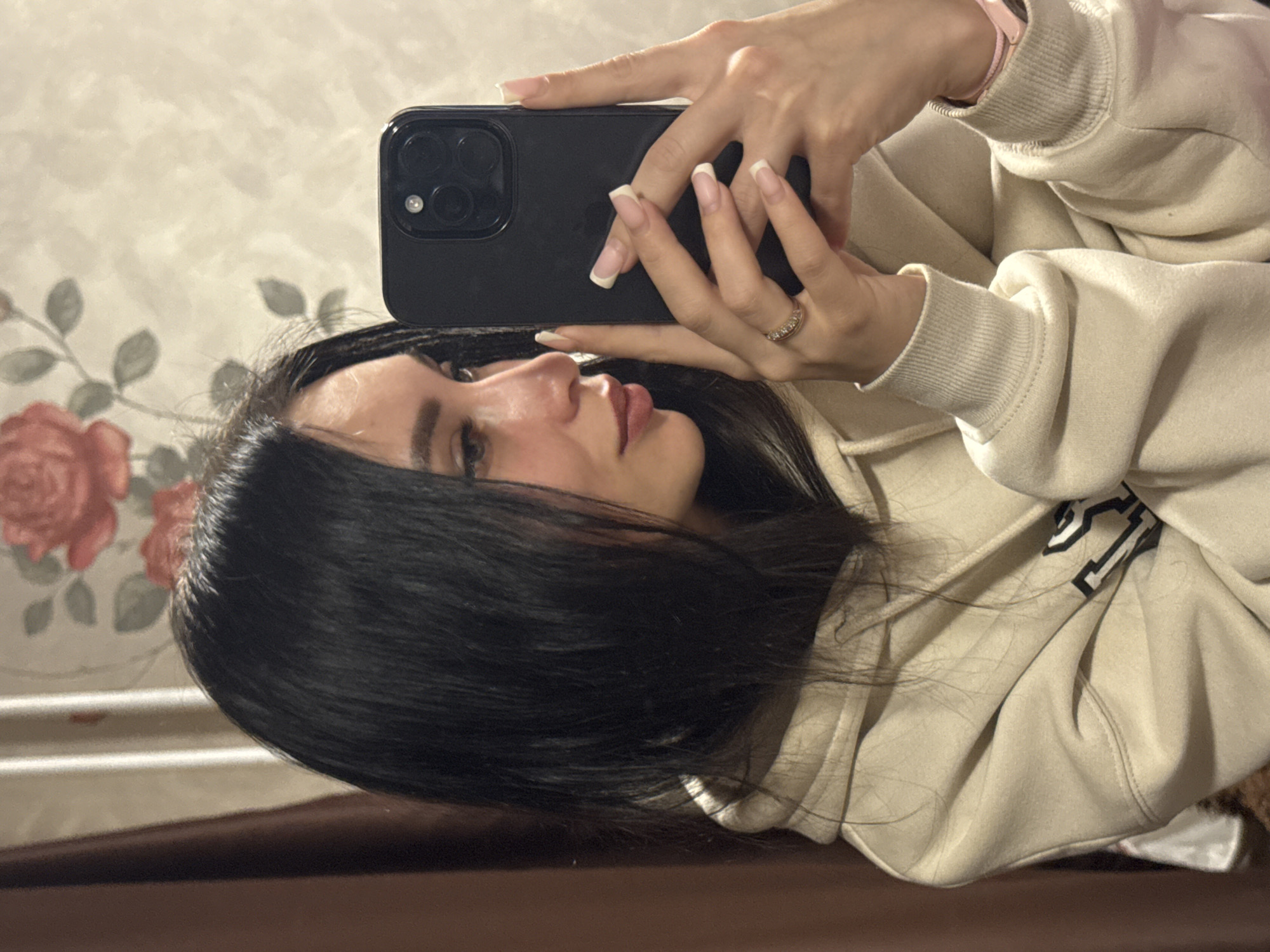 A young woman is shown taking a selfie indoors in Chaikovsky, Russia. The image is rotated 90 degrees clockwise, depicting her from a side profile as she holds a black smartphone, likely an iPhone, with both hands. Her gaze is directed towards the phone's screen. She has long, dark hair and is wearing a light-colored (beige or cream) hoodie with ribbed cuffs.
Key details include a ring with multiple small stones on her right ring finger and a pink wristband or watch on her left wrist. Her nails have a natural-colored base with white tips, resembling a French manicure. The background behind her features a wall with a floral pattern, including red roses and green leaves. Below the patterned wall, a dark, horizontal surface, possibly a wooden table or counter, is visible. The lighting suggests an indoor environment, and her expression appears neutral and focused on the task of taking the photo. No visible text can be clearly deciphered on her clothing, the phone, or in the background.