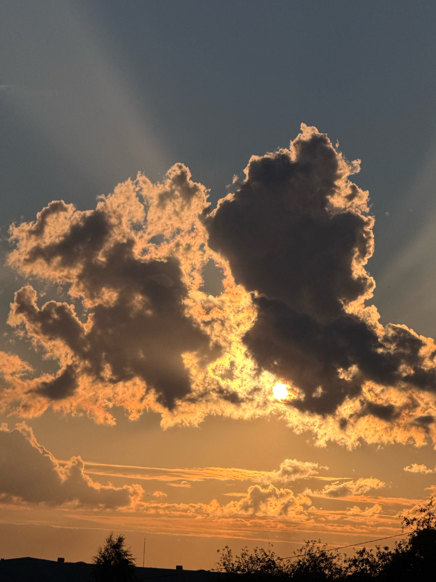 The image captures a dramatic sunset over the city of Galinė, Lithuania. The sky is a gradient of deep blue at the top transitioning to a warm, golden orange near the horizon. A large, dark cloud formation dominates the upper center of the frame, backlit by the setting sun, which is partially obscured behind it. Rays of light emanate from behind this cloud, creating a crepuscular effect. Below this, a band of thinner, more spread-out clouds are illuminated in shades of orange and yellow. The bottom of the image shows a silhouetted line of buildings and trees against the brightly colored sky, indicating a low urban or suburban landscape. The overall impression is one of a serene yet powerful natural phenomenon.