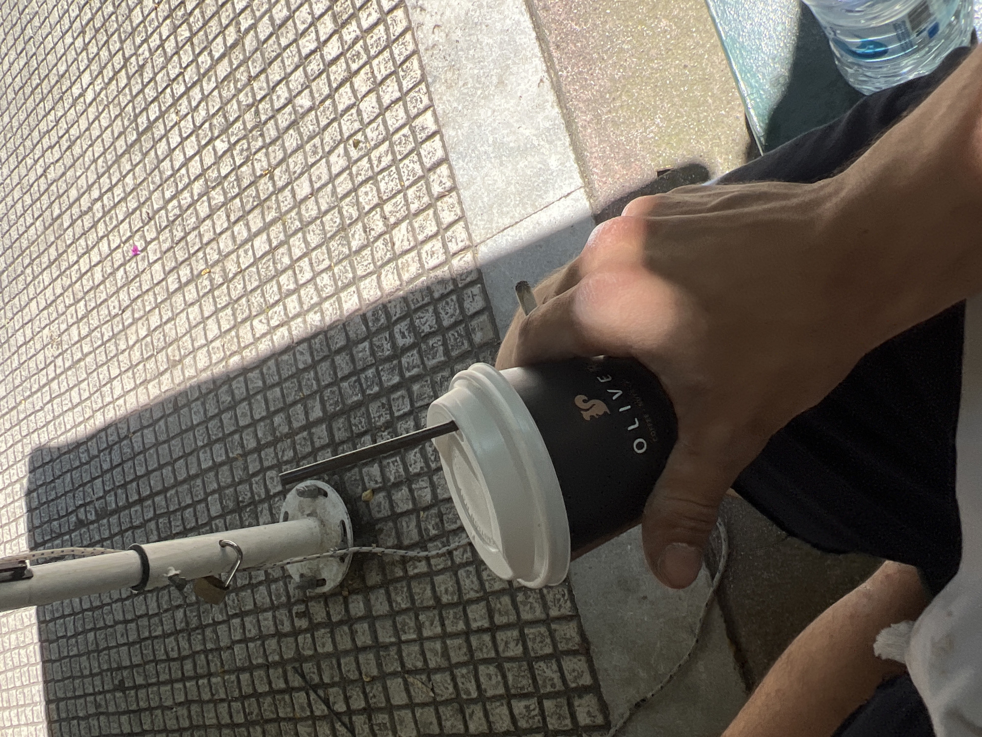 A person's hand, holding a black disposable coffee cup with a white lid and a dark straw, is visible in the foreground. The cup has a logo that includes a squirrel and the text "OLIVE COFFEE & NUTS". The person's fingers are wrapped around the cup, and another finger appears to be holding a cigarette. The person is wearing dark pants. In the background, there is a white pole with a black strap and a padlock, which is likely part of a street umbrella. The ground is paved with square tiles, and sunlight casts a shadow from the umbrella pole. The scene appears to be an outdoor seating area, possibly a cafe, in Athens, Greece.