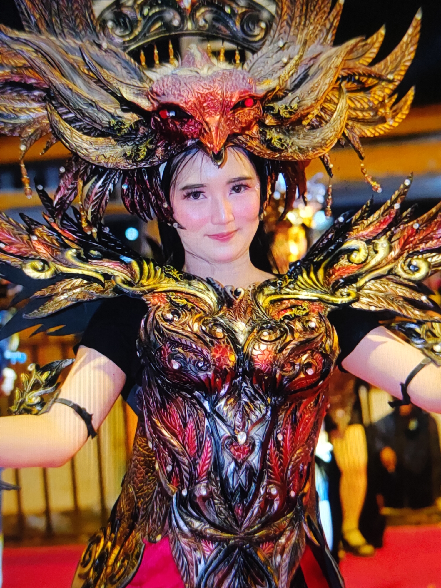 A medium shot captures a woman from the chest up, wearing an elaborate costume adorned with red, gold, and black feathery or scaled accents. The costume features a prominent headdress resembling a bird of prey with glowing red eyes, and ornate pauldrons that extend outwards. The woman has fair skin, dark hair, and is smiling faintly, looking towards the camera. Her makeup is subtle, with blush on her cheeks. The background is blurred, showing some indistinct lights and figures, suggesting an outdoor or dimly lit indoor event. The overall impression is that of a performer in a festival or parade. The location is unknown, Unknown.