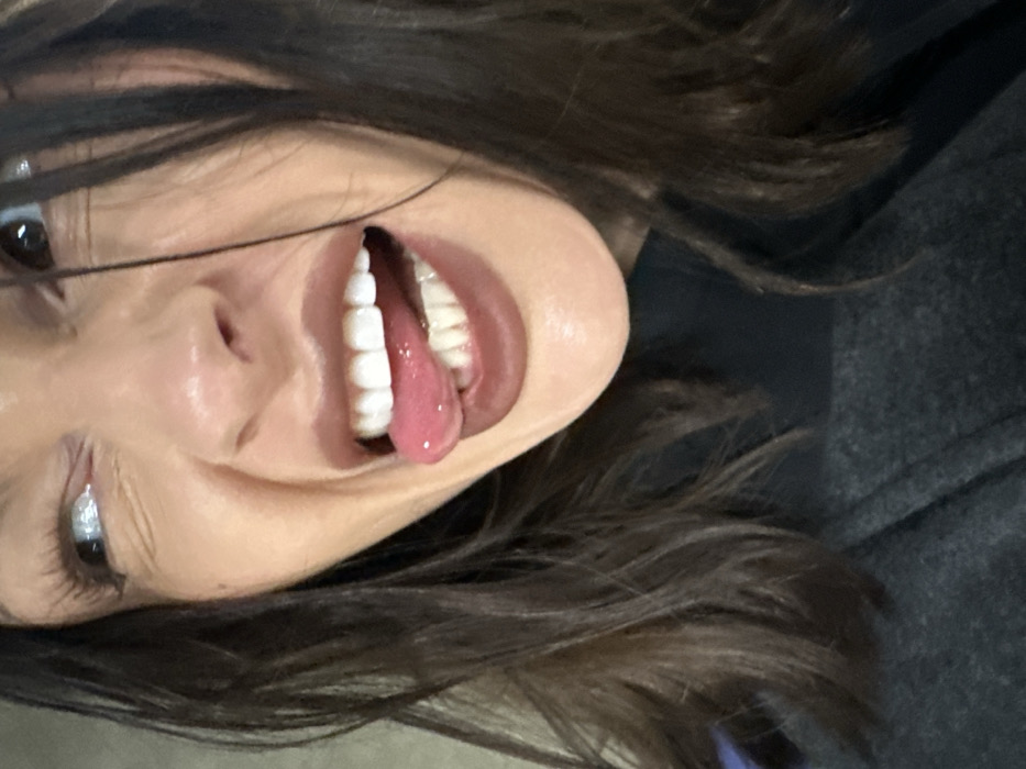 The image displays a close-up, cropped portrait of a female individual from the upper torso upwards. She has dark, wavy hair framing her face. Her head is slightly tilted, and she is making an expressive facial gesture. Her mouth is open in a wide grin, revealing both upper and lower white teeth. Her pink tongue is extended, slightly curled, and rests on her lower front teeth. She wears dark brownish-red lipstick. Her left eye, visible in the upper right quadrant of the image, looks upwards and to the right, showing a significant portion of the white sclera. Her nose is slightly crinkled. She is dressed in a dark, high-necked garment under a darker, possibly charcoal grey, outer layer. A small section of purple fabric is visible on the far left edge of the frame, near her shoulder. The background is uniformly light, out of focus, and featureless, appearing as a plain wall or surface. The environment is nondescript.