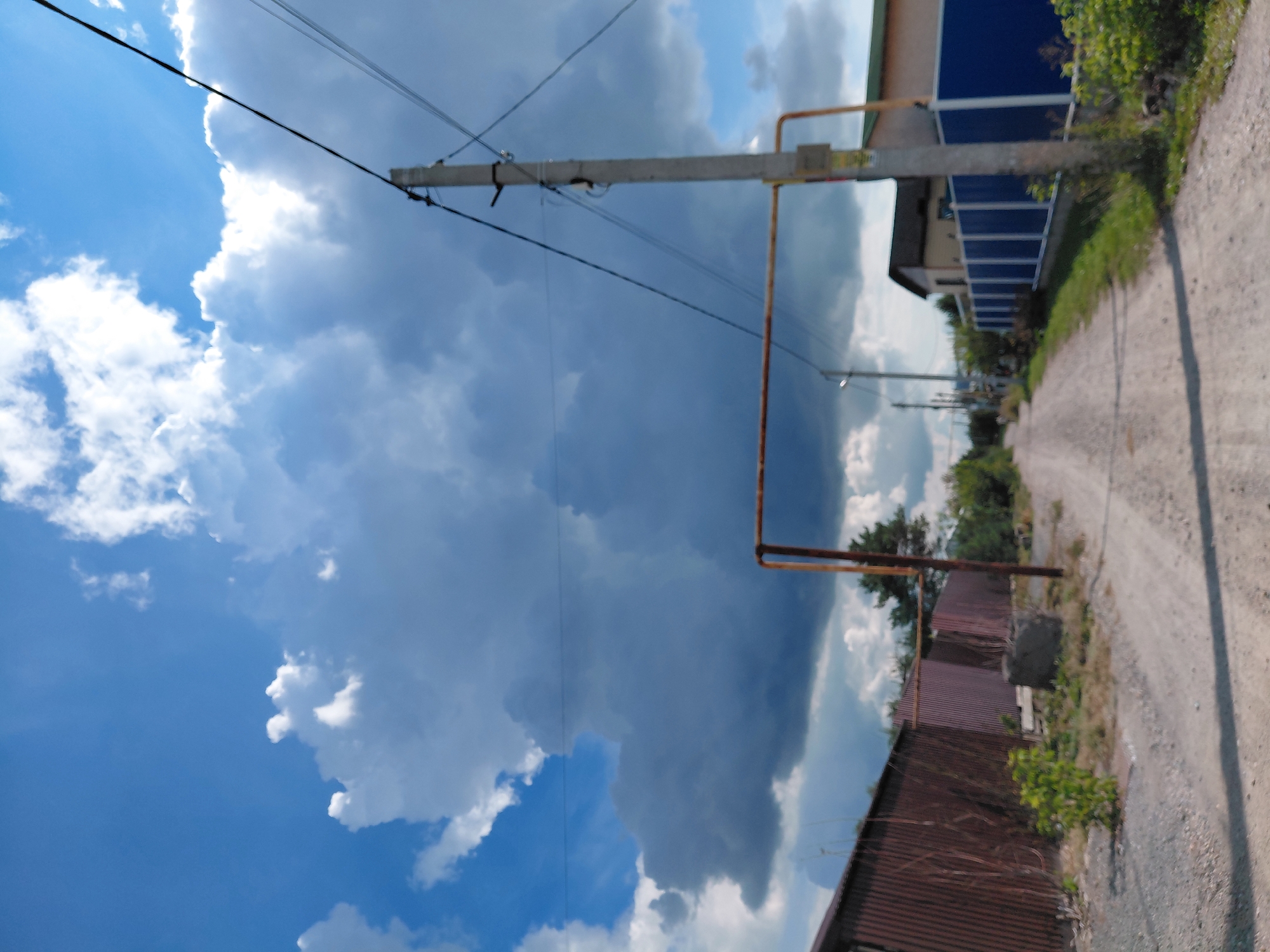 The image depicts a rural or suburban dirt road in Novosibirsk, Russia, on a partly cloudy day. The road is unpaved and appears to be a typical unmaintained track, with gravel and dirt. On the left side of the road, there is a long, rusty corrugated metal fence or building. To the right, there is a concrete utility pole with electrical wires running across the frame. Further down the road on the right, a modern house with a dark roof and a blue fence is visible. Tall green bushes and trees line the sides of the road and surround the properties. A prominent horizontal rusted metal pipe crosses the road in the middle distance, supported by utility poles. The sky is a mix of bright blue and large, dark, dramatic storm clouds. No people or vehicles are present in the scene. The overall impression is one of a quiet, possibly isolated location, under a dynamic sky.