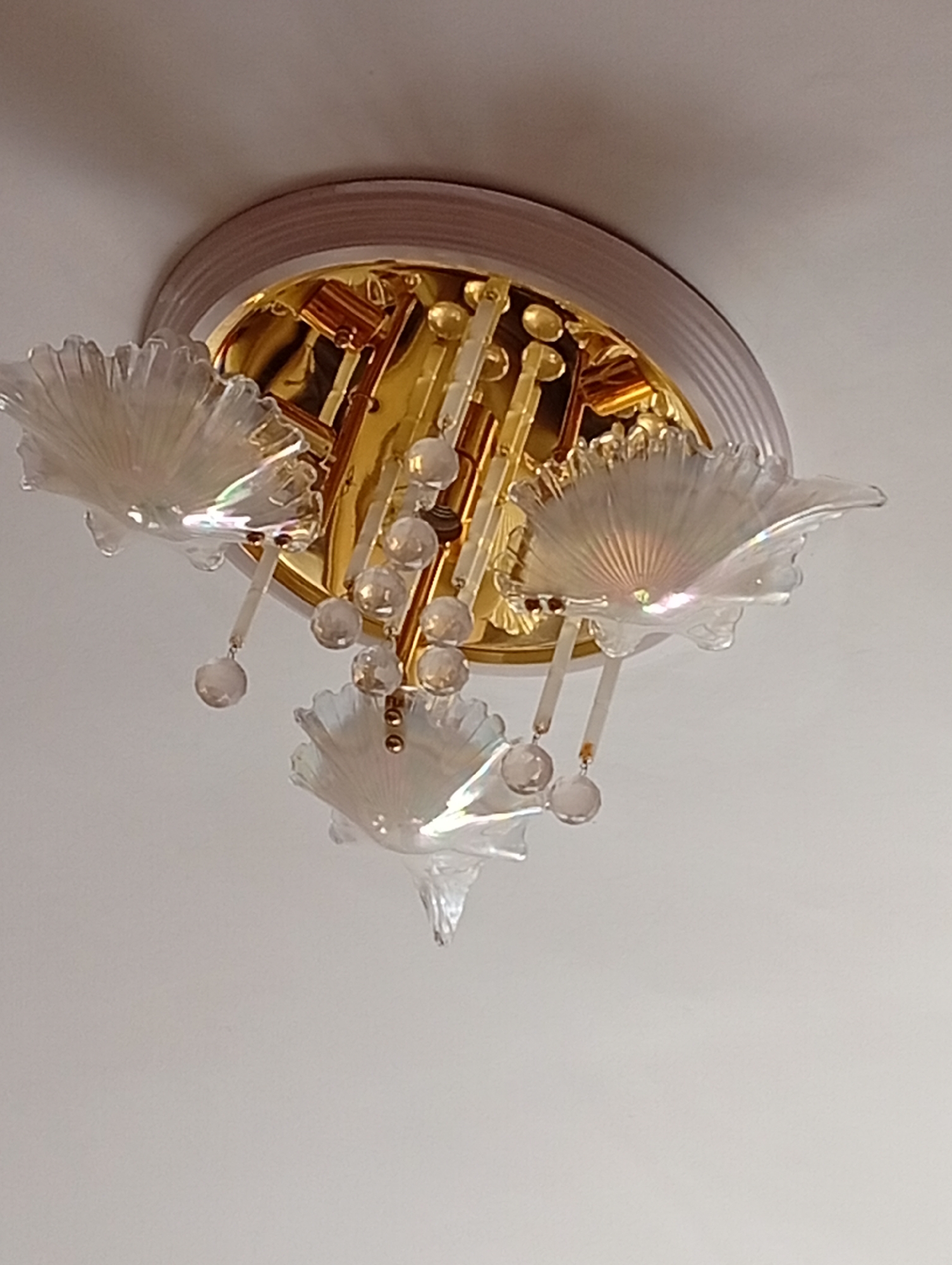 A close-up, low-angle shot shows a modern, ornate ceiling light fixture against a white ceiling. The fixture has a gold-toned base with clear, fan-shaped glass elements that resemble shells. Several clear glass beads hang from the fixture. The light fixture appears to be mounted on the ceiling of a room. There is no indication of the location being in Veliatyno, Ukraine, as the image provides no contextual clues to support this.