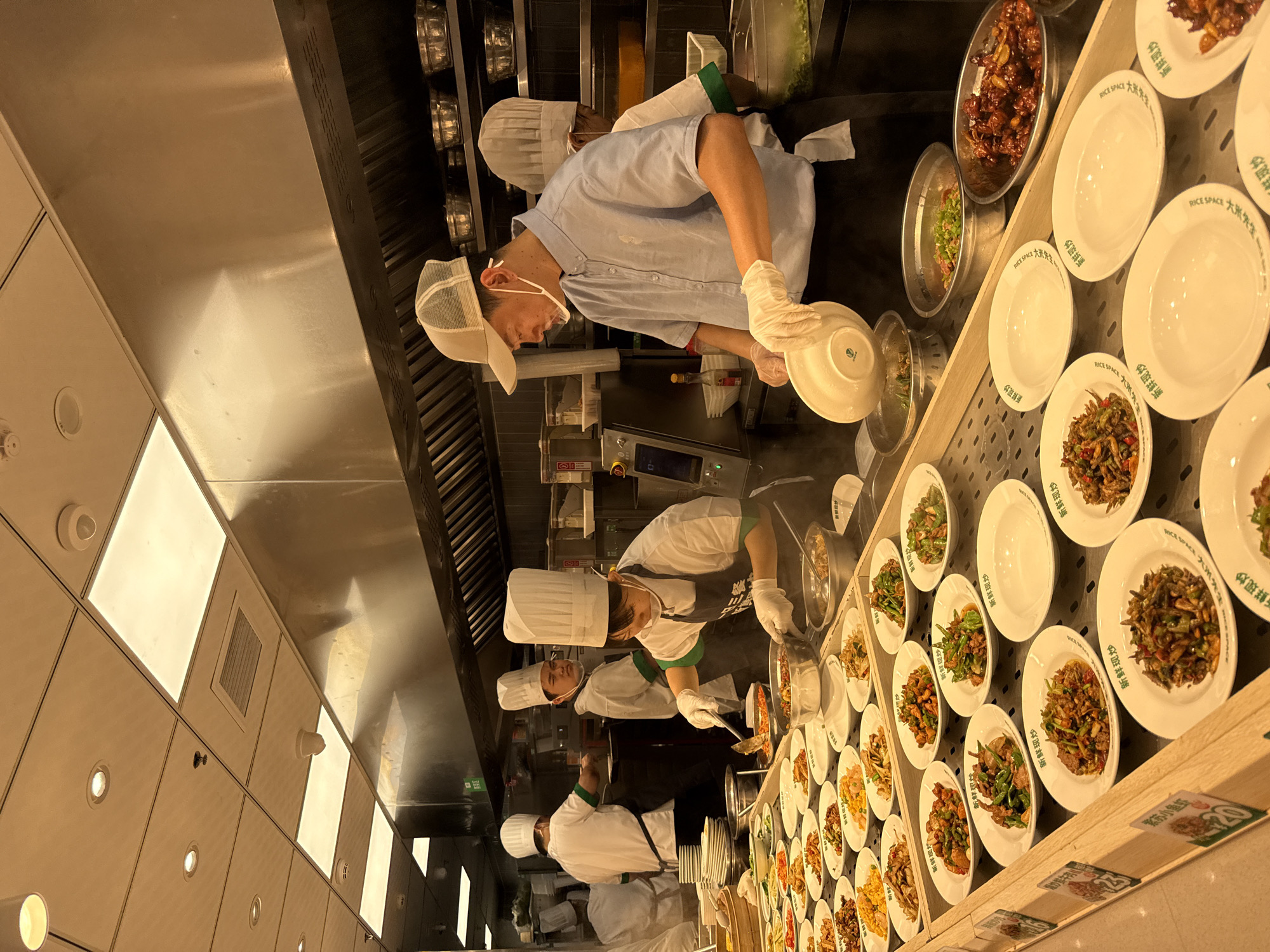 In a brightly lit, commercial kitchen in Tianhe District, China, four chefs in white toque hats and aprons are actively engaged in food preparation and service. The scene is dominated by stainless steel appliances and overhead ventilation hoods, indicative of a professional culinary environment. In the foreground, a chef wearing a light blue button-down shirt and a mesh-back cap with a transparent face shield is meticulously placing food into a white bowl held in their gloved hand. This chef is positioned behind a long counter displaying numerous white plates filled with various prepared dishes, appearing to be a self-service or cafeteria-style food station.
To the left, another chef in a white toque and apron is using tongs to serve food from a large pot into a bowl. Further back, two other chefs in similar attire are also visible, one appearing to be assisting with food preparation and the other standing by. The counter is laden with an array of colorful food items in individual servings, suggesting a buffet or grab-and-go setup. The overall atmosphere is one of organized culinary activity within a well-equipped kitchen.